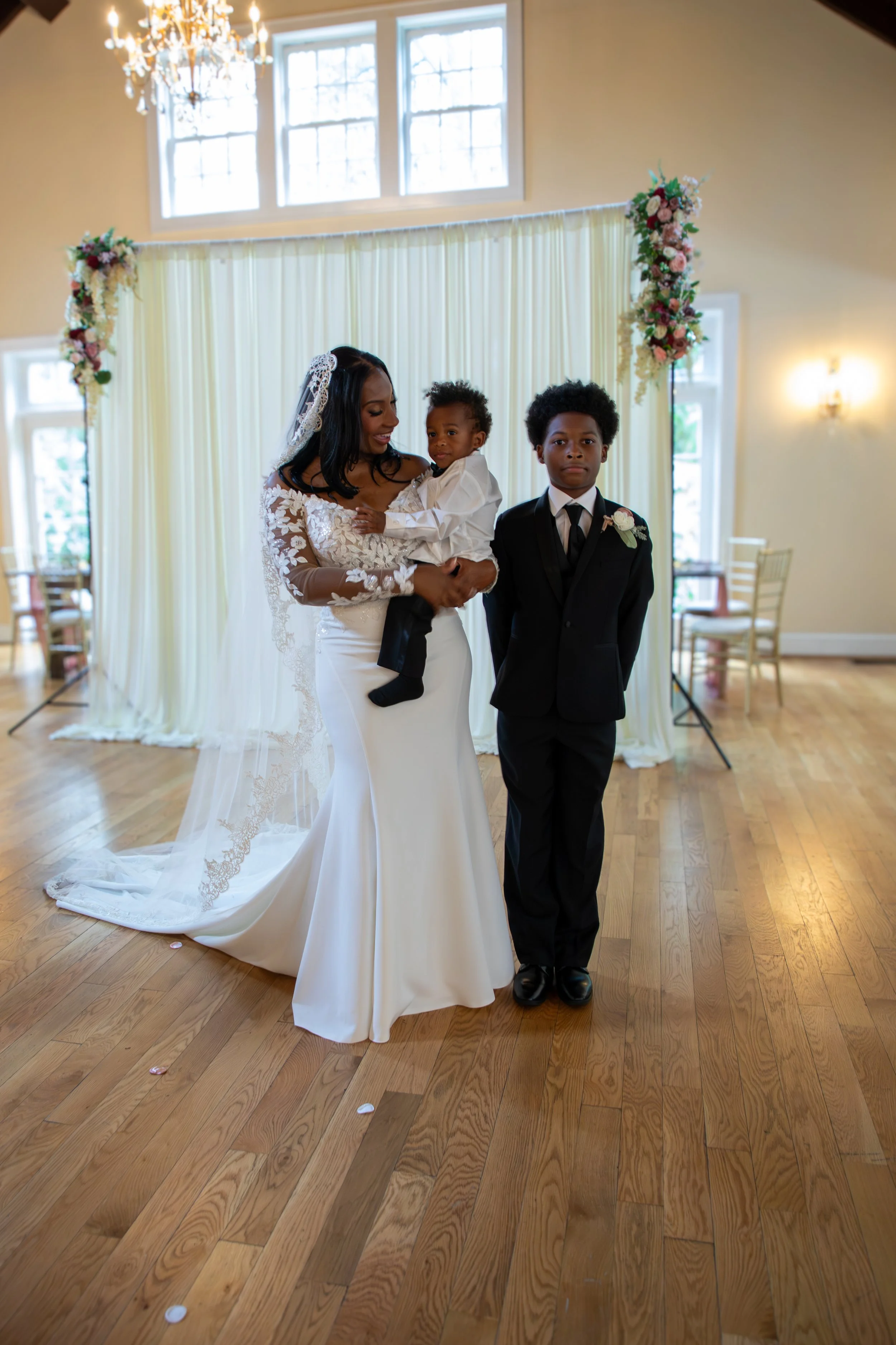 Photo & Video by Will Locke Wedding Photography at the Woman's Club of Portsmouth in VA. A bride in a white wedding gown holding a young child, standing next to a boy in a black suit, during a wedding ceremony in a decorated indoor venue.