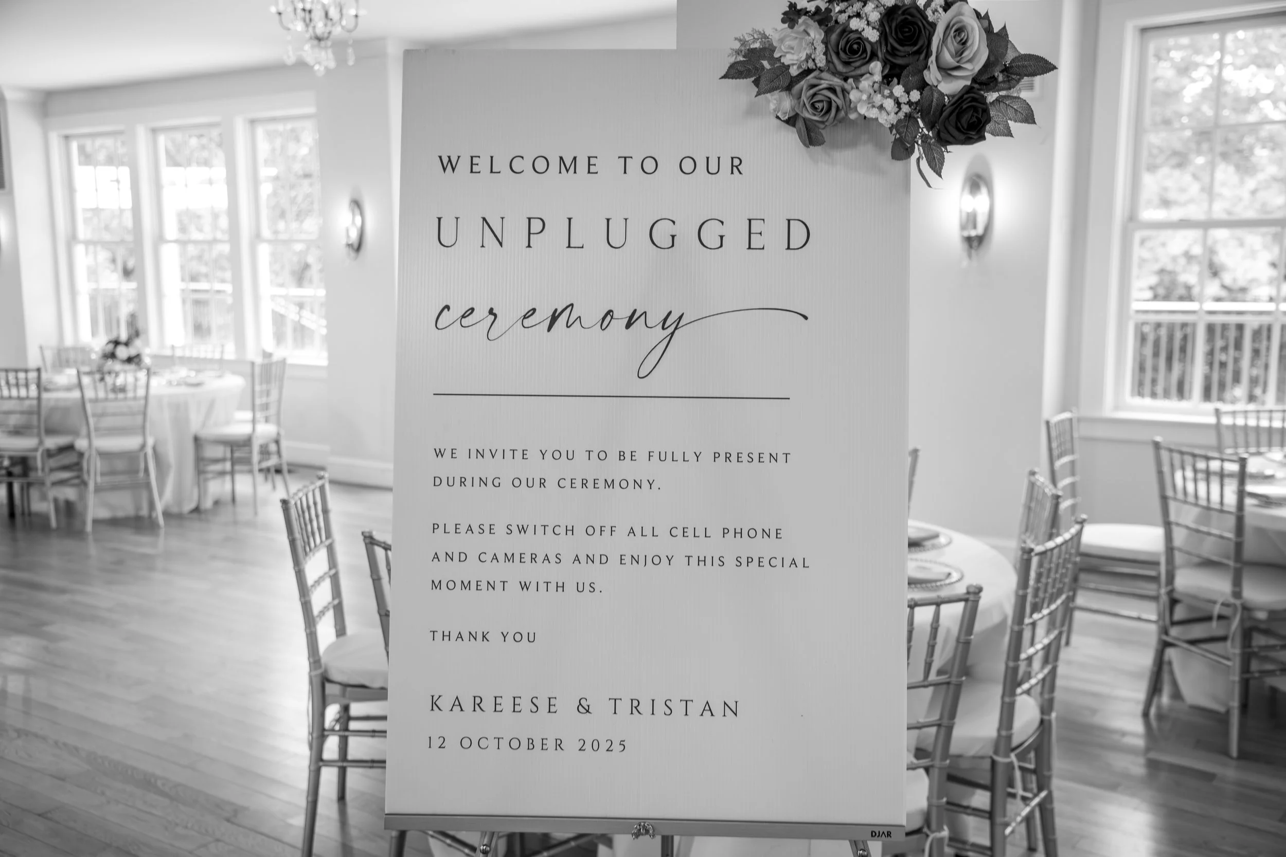 Wedding Photography by Will Locke. A wedding reception venue with a white welcome sign on an easel, decorated with a bouquet of roses in shades of pink, purple, and white, in a bright room with large windows, elegant chandeliers, and gold chairs.