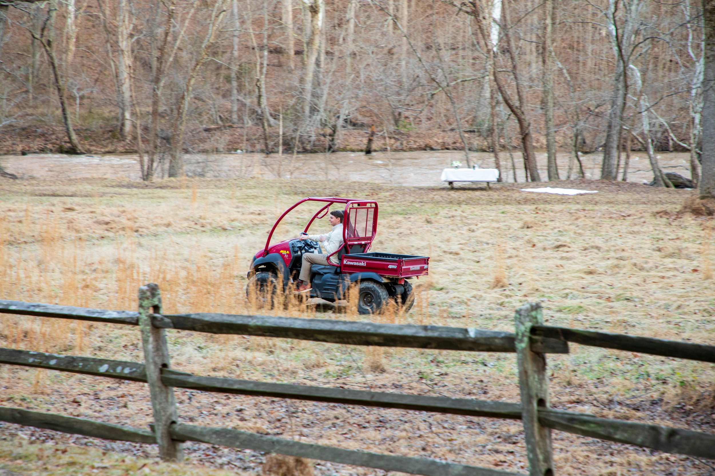 Engagement Photography by Will Locke near Richmond, VA in Montpelier. A person driving a red Kawasaki utility terrain vehicle (UTV) across a grassy field near a wooden fence and a wooded area.