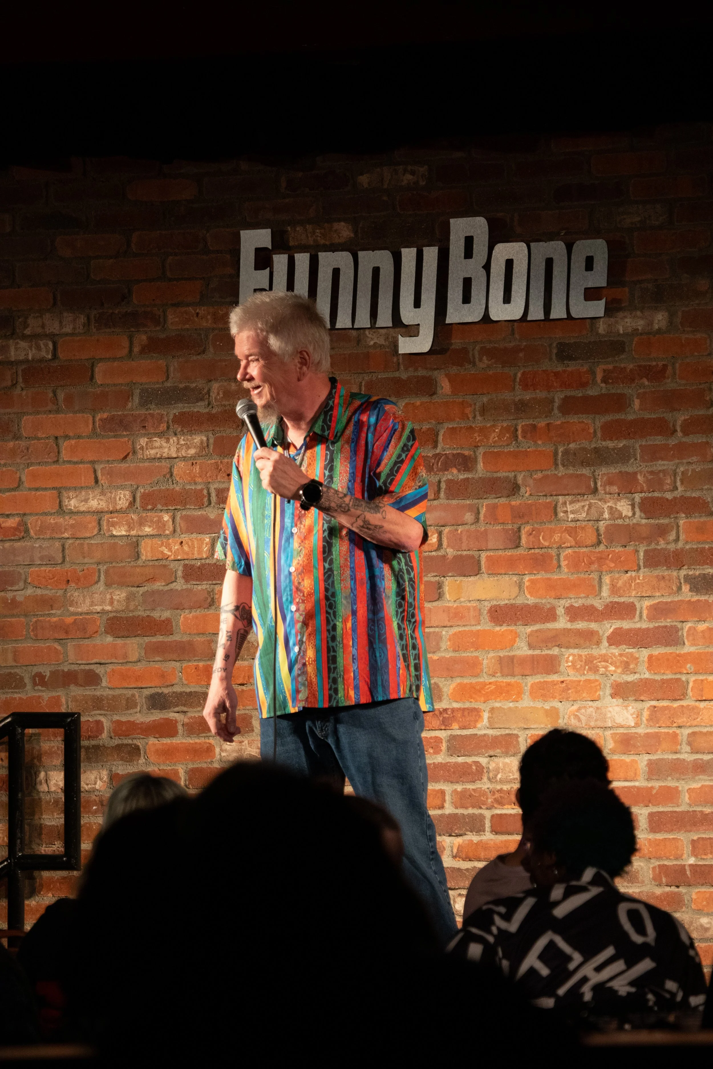 Comedy Photography by Will Locke. A comedian performing on stage at Funny Bone comedy club with brick wall background, wearing a colorful striped shirt and holding a microphone