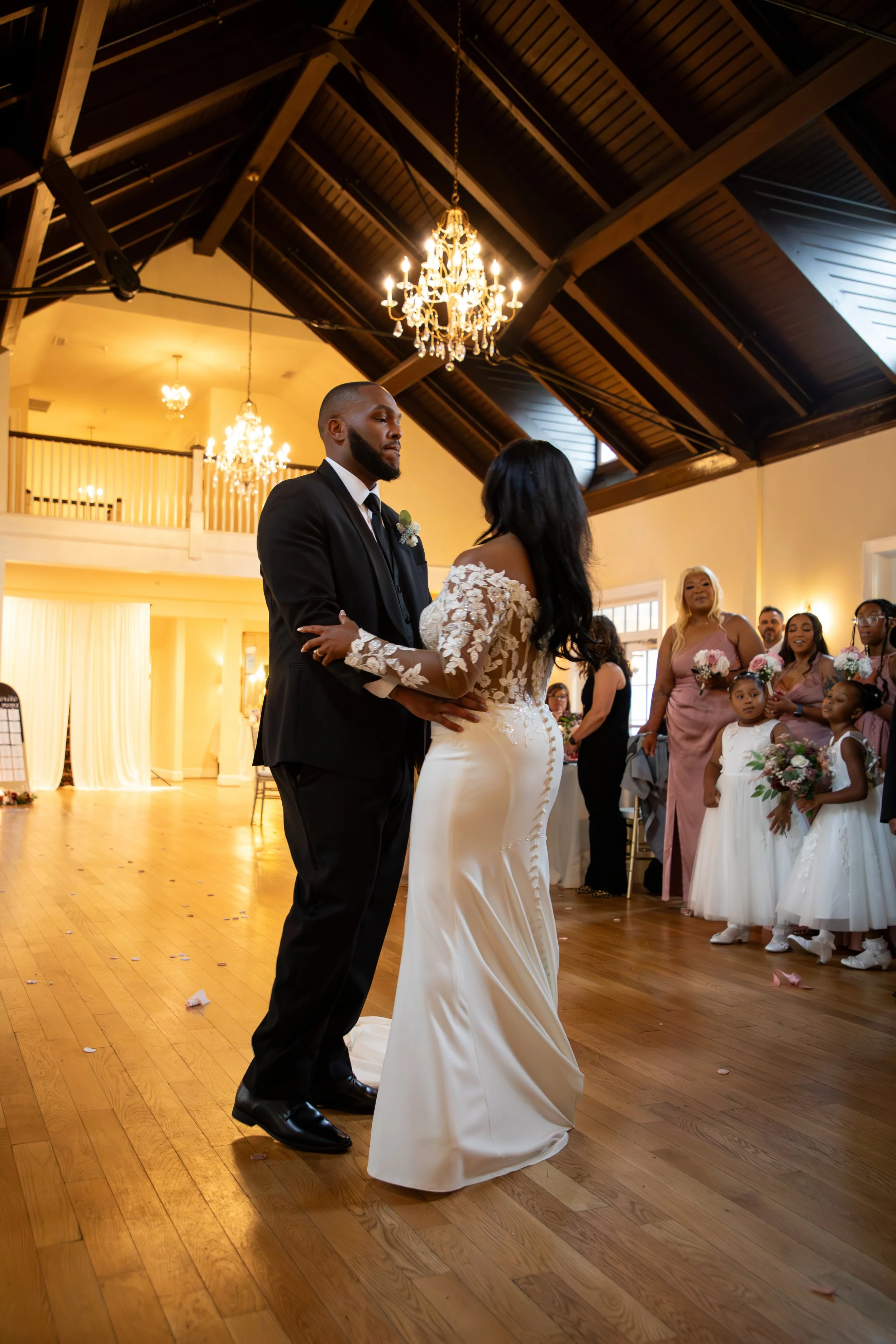 Wedding Photography by Will Locke. A bride and groom dancing during their wedding reception, surrounded by bridesmaids and flower girls, in a decorated indoor venue with chandeliers.