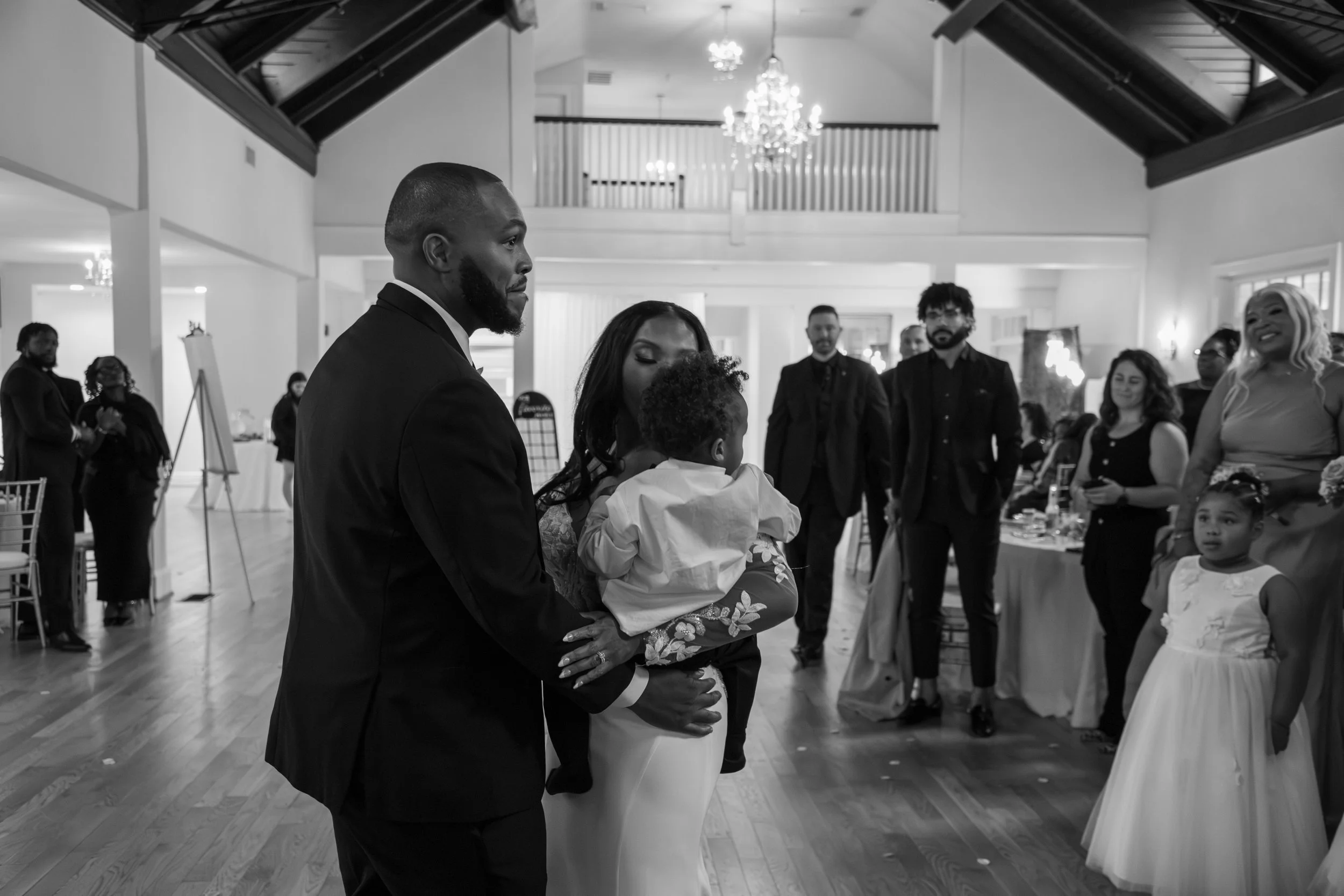 Photo & Video by Will Locke Wedding Photography. A black and white photo of a wedding reception, with a man in a black suit holding a small child and a woman in a dress standing nearby, surrounded by onlookers in formal attire in a decorated ballroom
