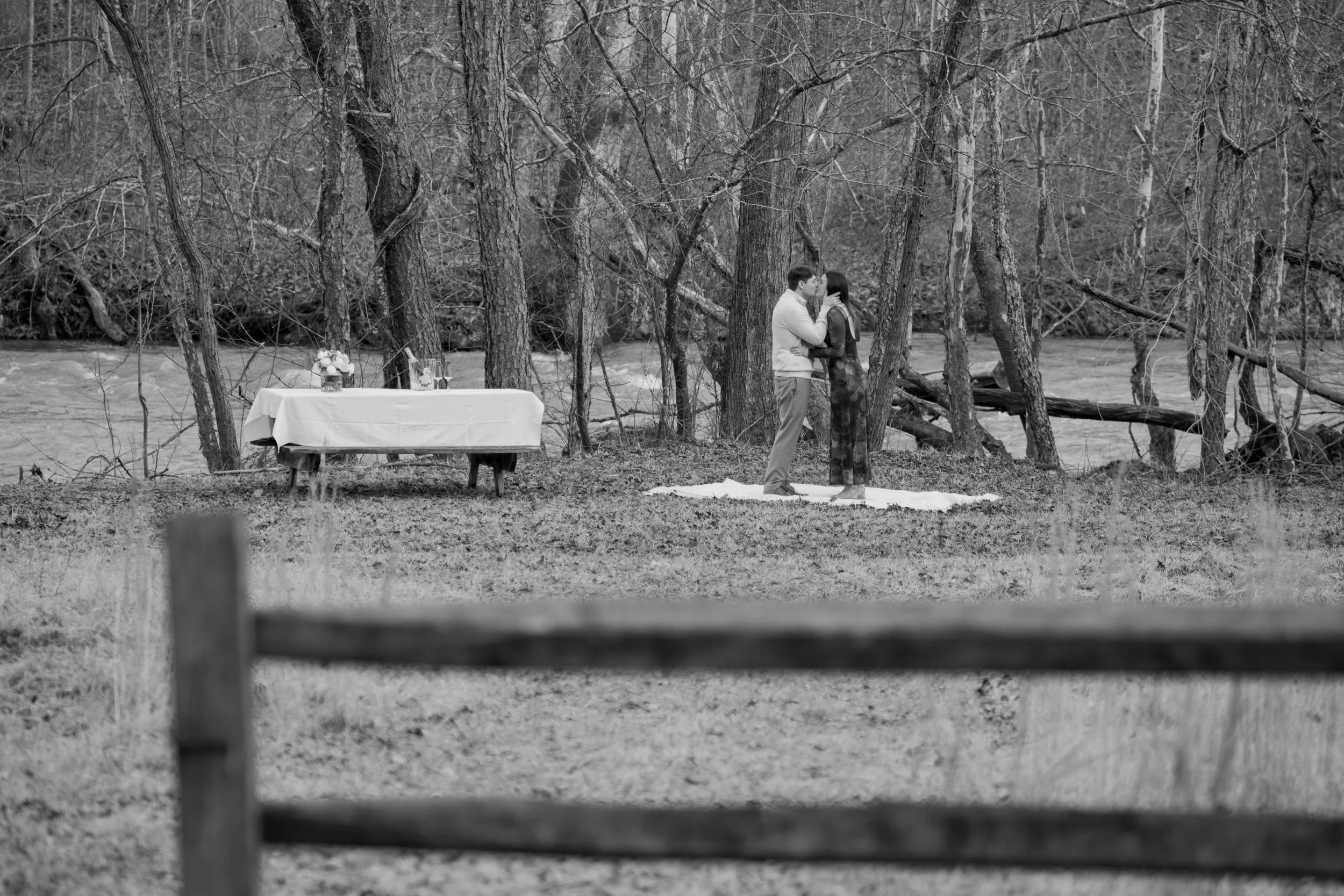 Engagement Photography by Will Locke near Richmond, VA in Montpelier. A couple hugging each other outdoors near a river, standing on a white blanket on the ground, surrounded by leafless trees in a forested area.
