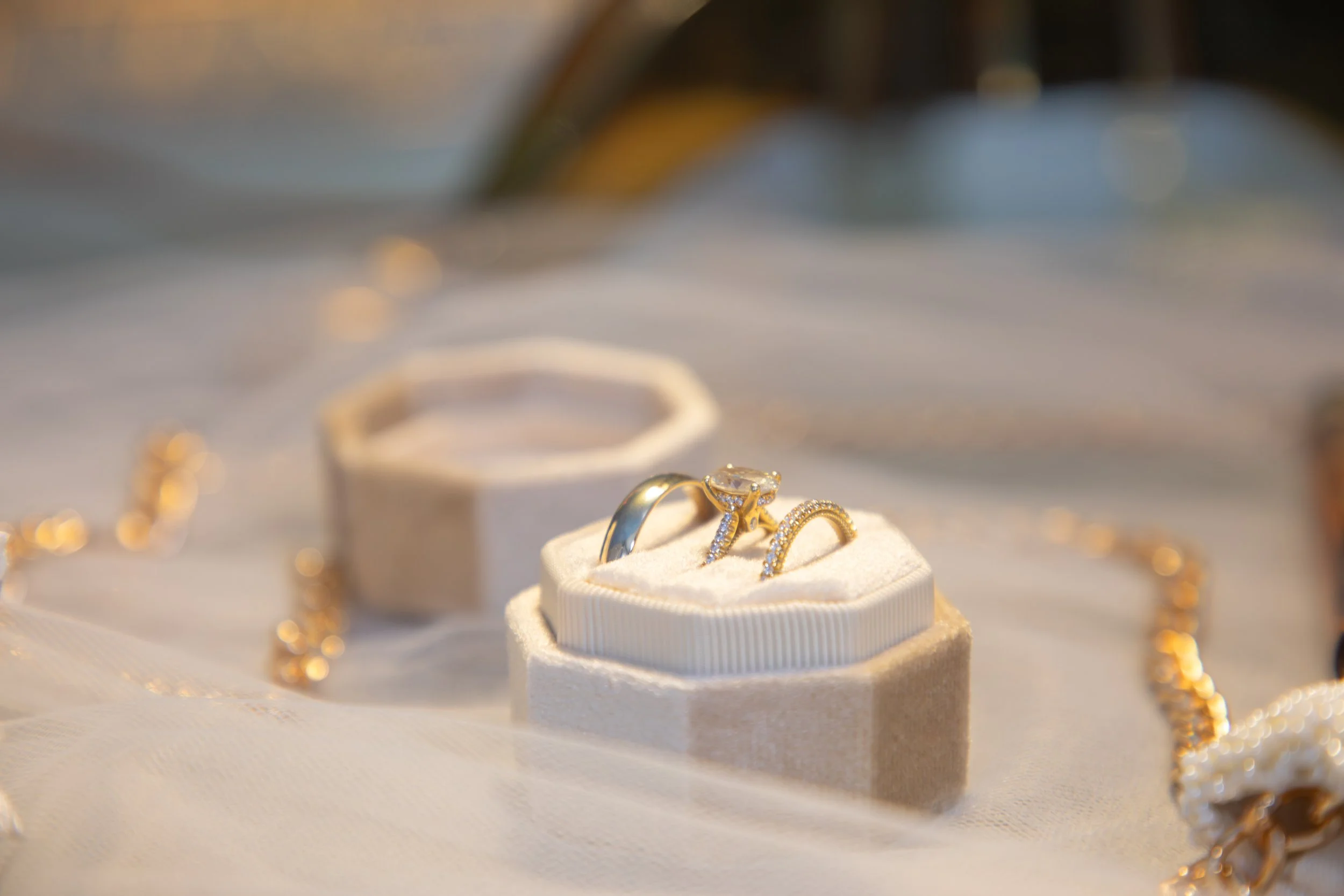 Wedding Photography by Will Locke. Close-up of a jewelry display with wedding and engagement rings on a white cushion, surrounded by gold chains on a soft, white fabric surface.