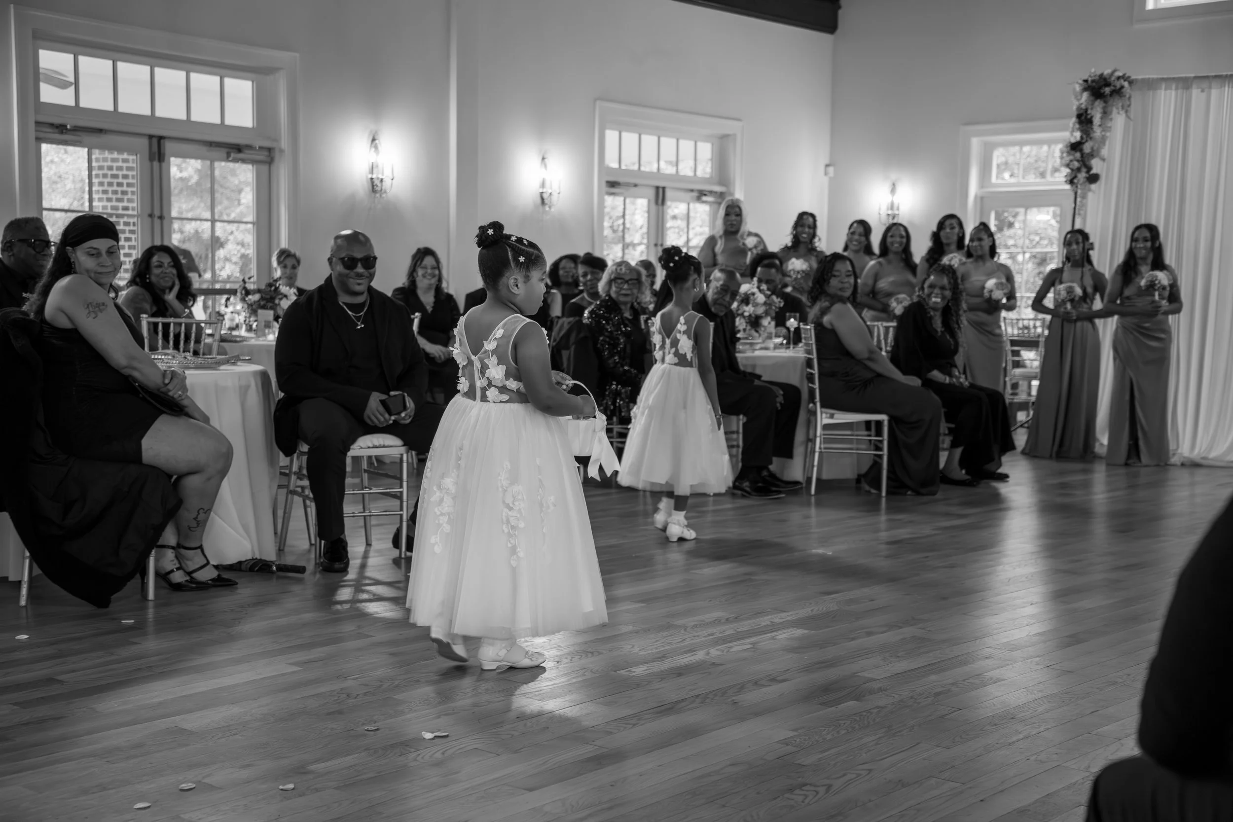 Wedding Photography by Will Locke. A wedding reception with two flower girls walking across the dance floor, surrounded by seated guests and bridesmaids standing along the wall, in a well-lit room with large windows and floral decorations.