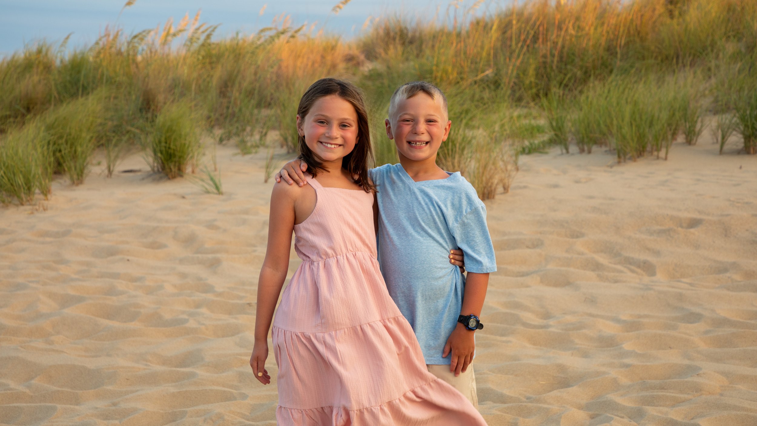 Photo & Video by Will Locke Family Photography in Virginia Beach, VA. Two children, a girl in a pink dress and a boy in a blue shirt, standing on a sandy beach with dune grass in the background, smiling at the camera.