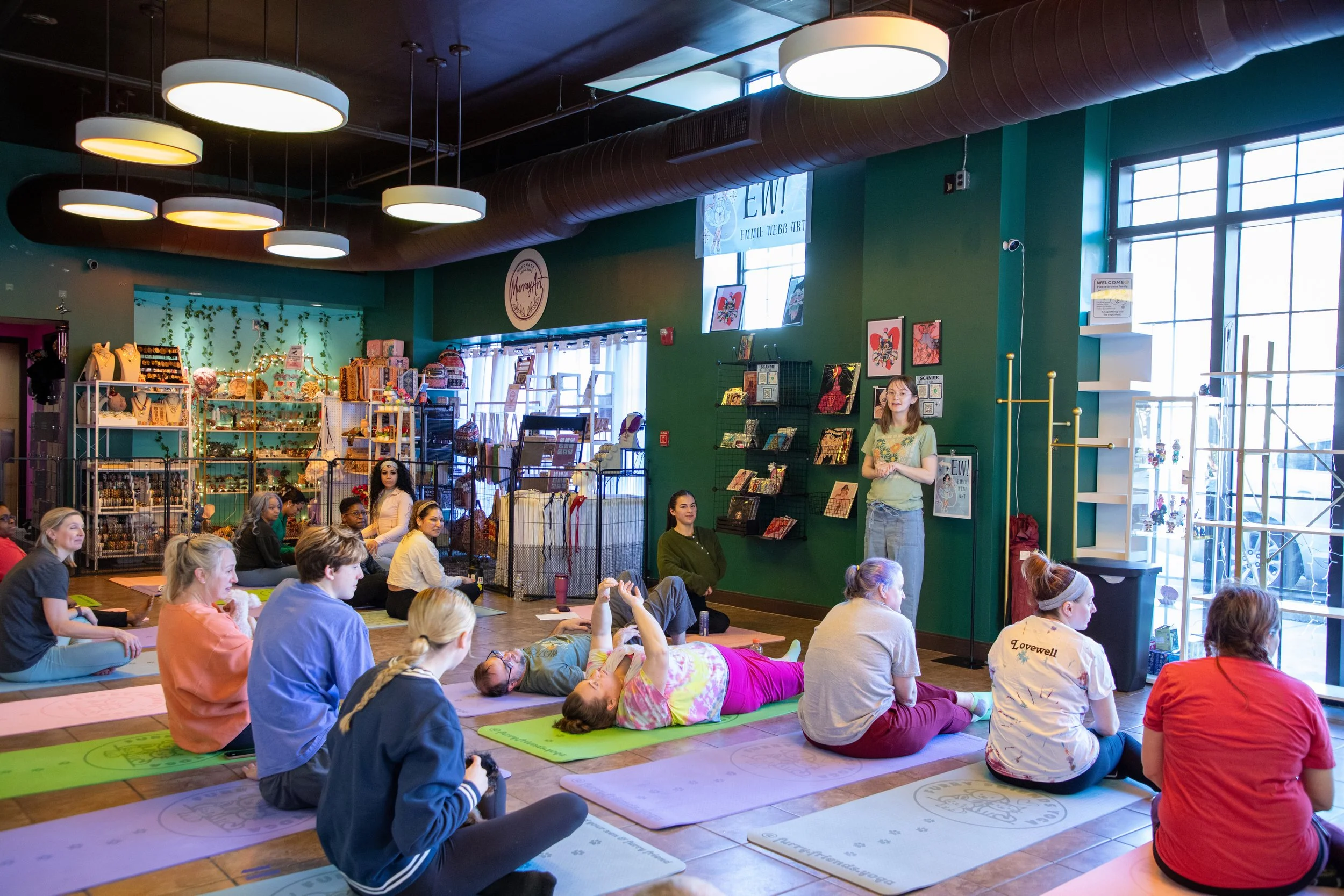 A yoga class in session inside a retail store. Participants are seated or lying on yoga mats, facing a female instructor standing in front of a green wall with artwork and books. Marketing and Event Photography by Will Locke. 