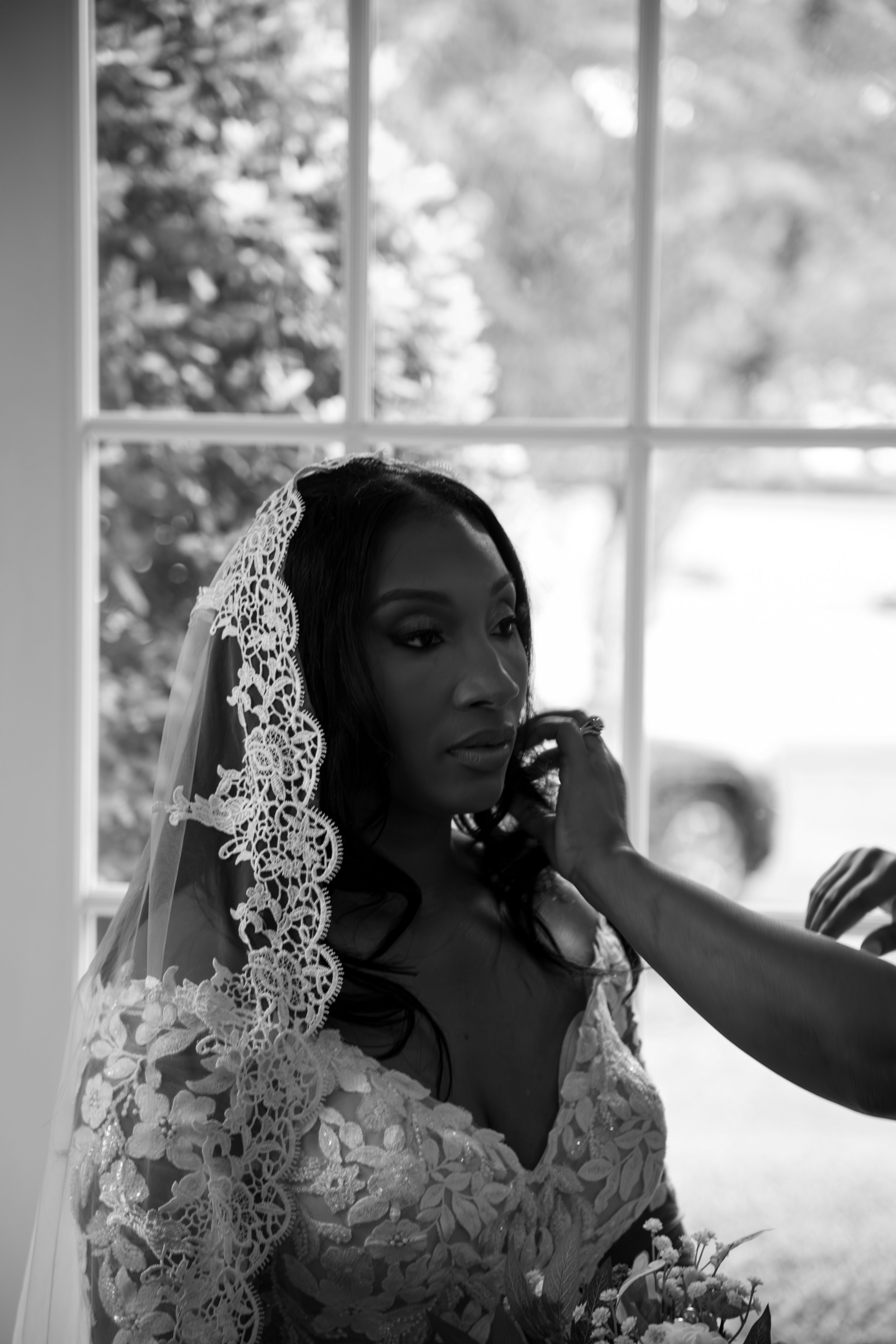 Wedding Photography by Will Locke. Black and white photo of a woman in a wedding dress with lace details, sitting by a window while someone adjusts her hair or makeup.