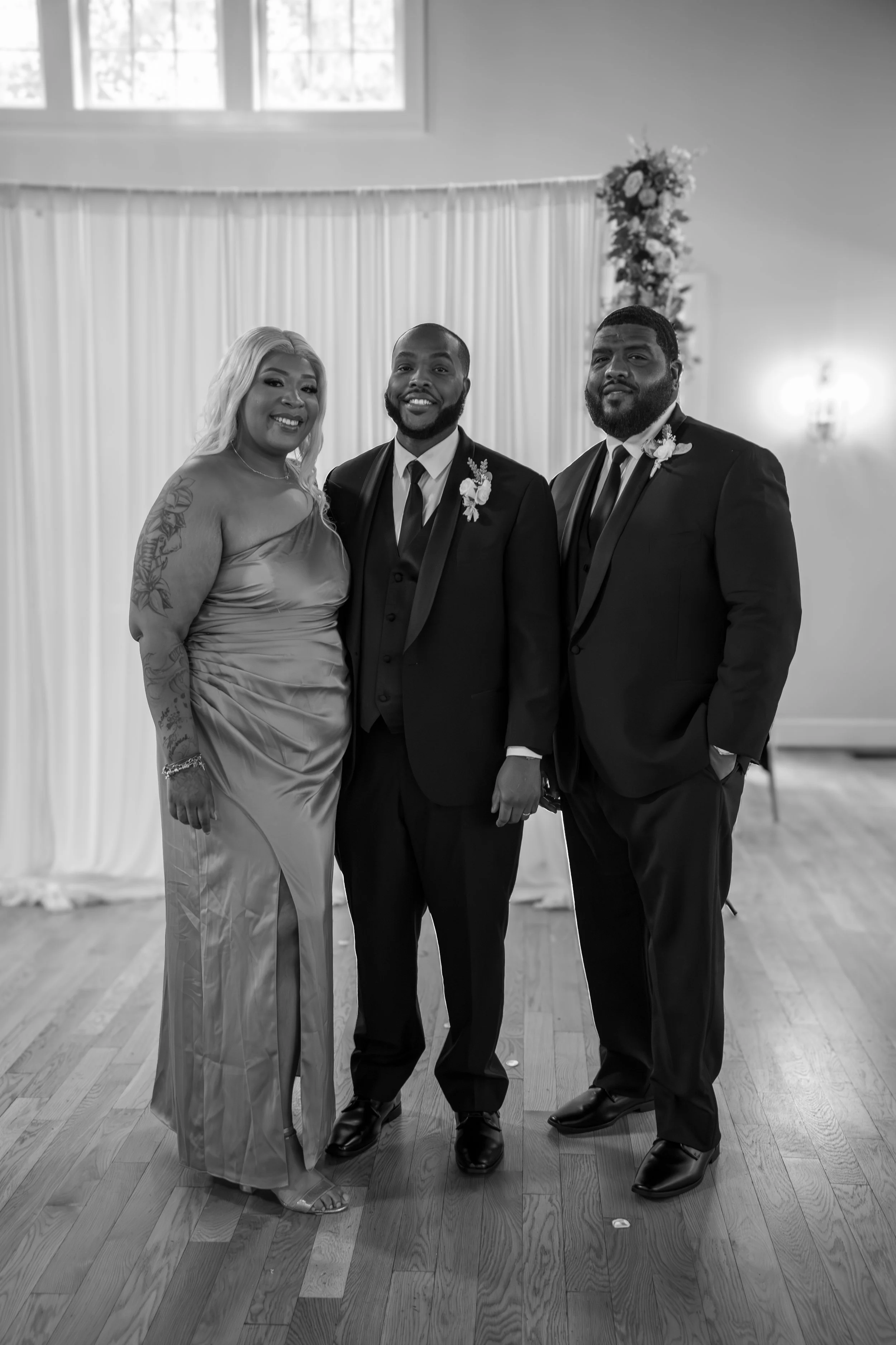 Photo & Video by Will Locke Wedding Photography at the Woman's Club of Portsmouth in Virginia. A black-and-white photo of three people dressed in formal attire, standing together indoors with a curtain and floral arrangement in the background.