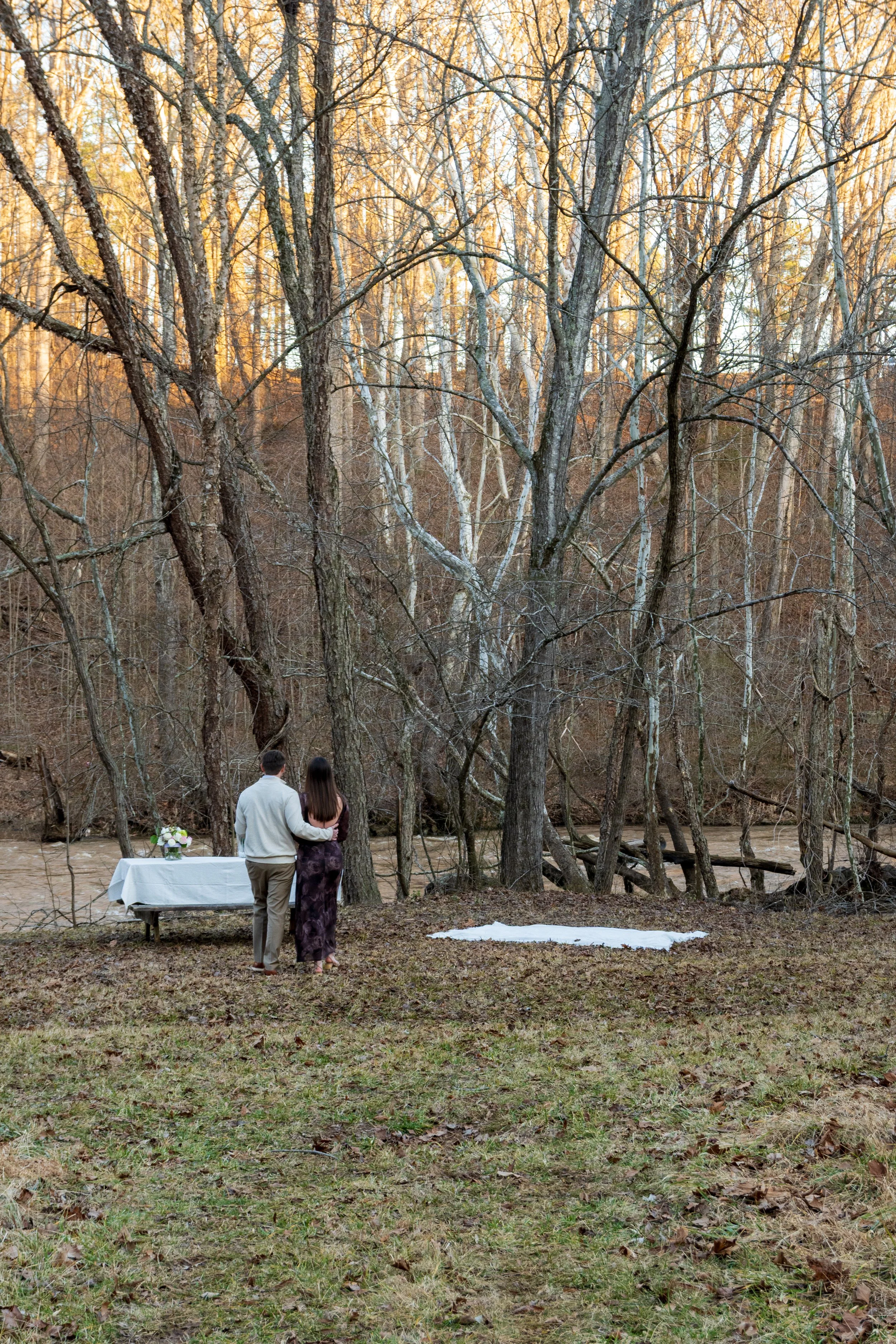 Engagement Photography by Will Locke near Richmond, VA in Montpelier. A couple walks to a white cloth on the ground near a wooded area with a stream on a fall day, with a table on the left side with white tablecloth.