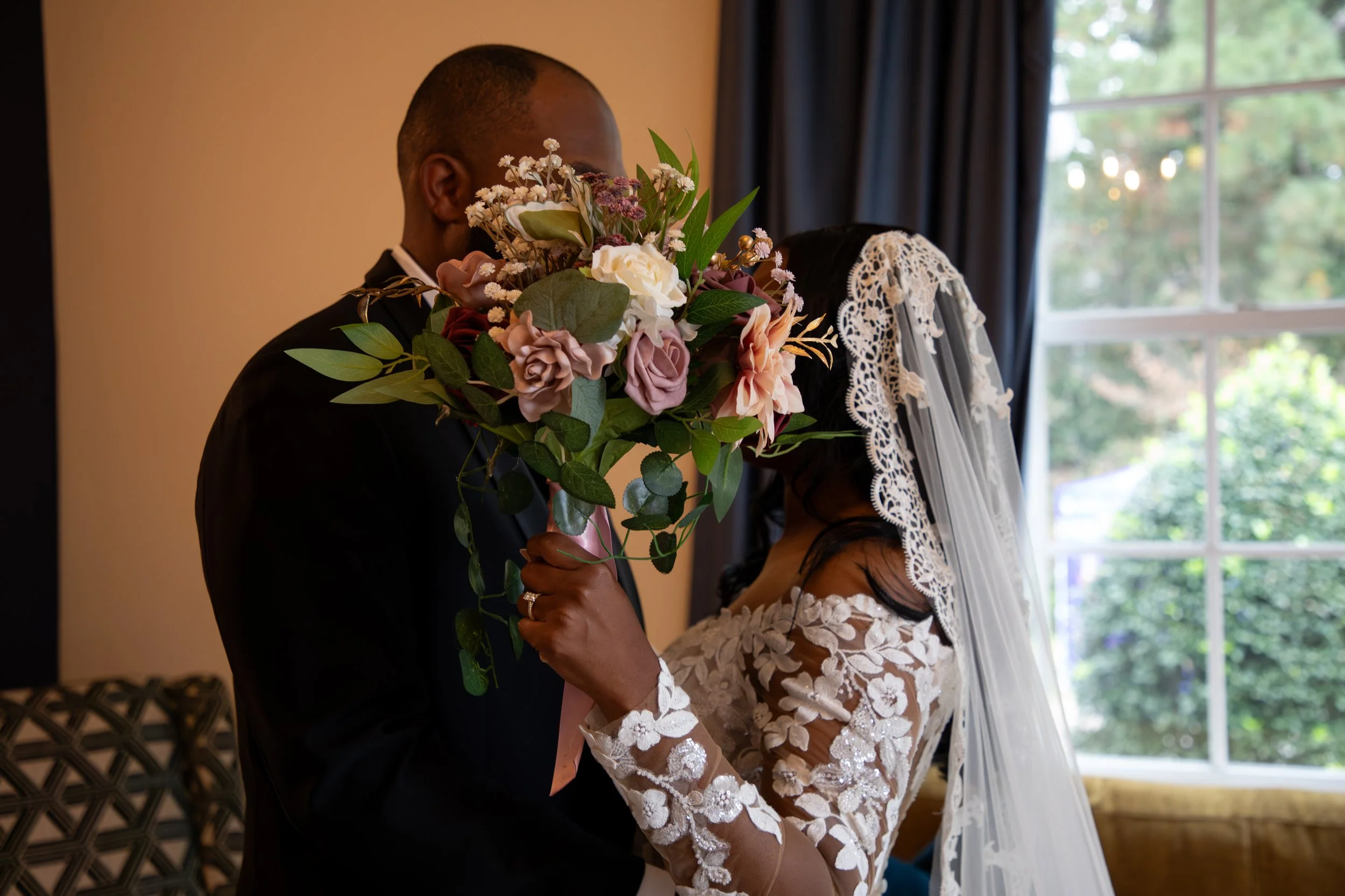Wedding Photography by Will Locke. A wedding scene with a bride holding a bouquet of pink, white, and purple flowers and a groom in a black suit, both standing near a window with a lace veil covering part of the bride's face.