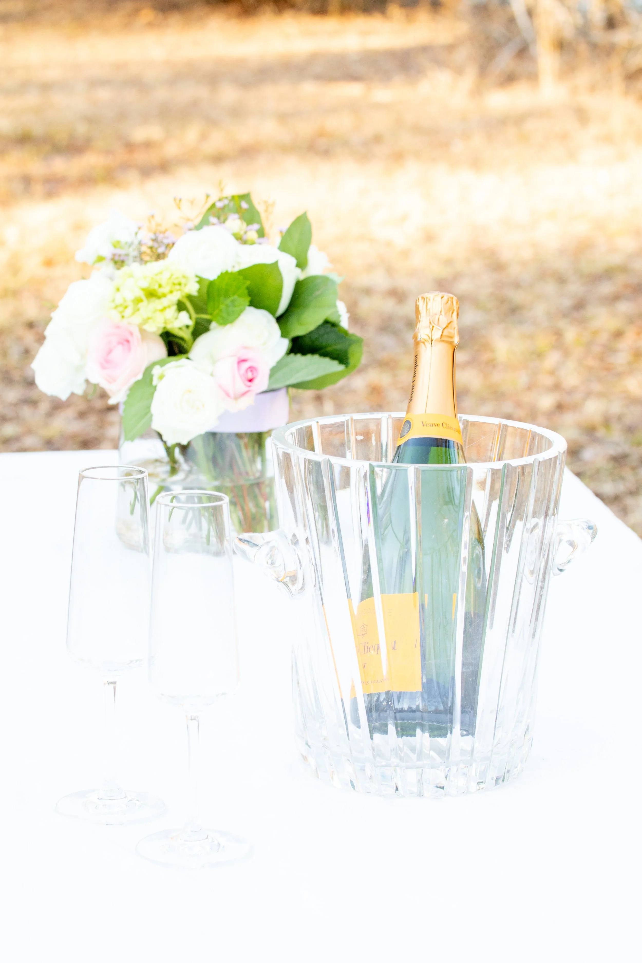 Engagement Photography by Will Locke near Richmond, VA in Montpelier. A bottle of Veuve Clicquot champagne in an ice bucket with two empty champagne flutes on a white table outdoors, with a flower arrangement. 