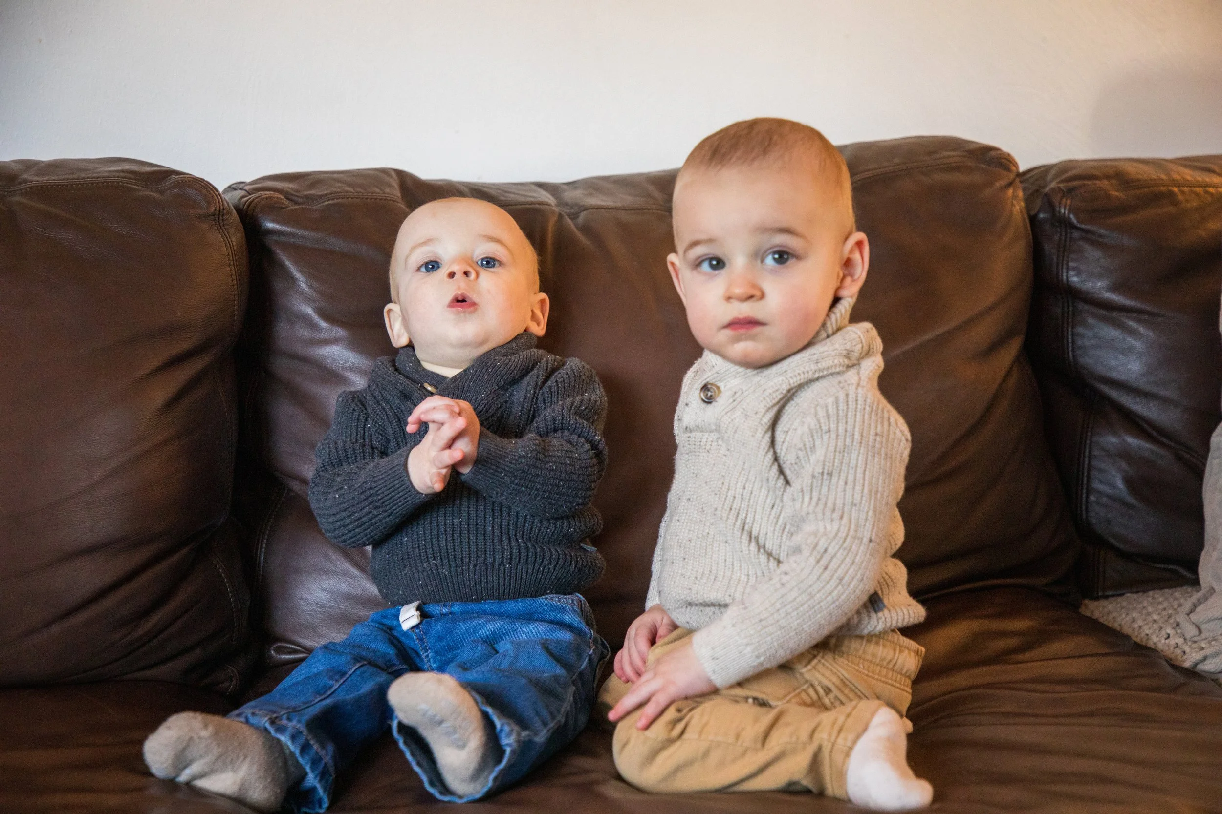 Newborn Baby Photography by Will Locke. Two young boys sitting on a brown leather couch, one wearing a dark gray sweater and blue jeans with socks, the other wearing a beige sweater and tan pants, both looking at the camera.