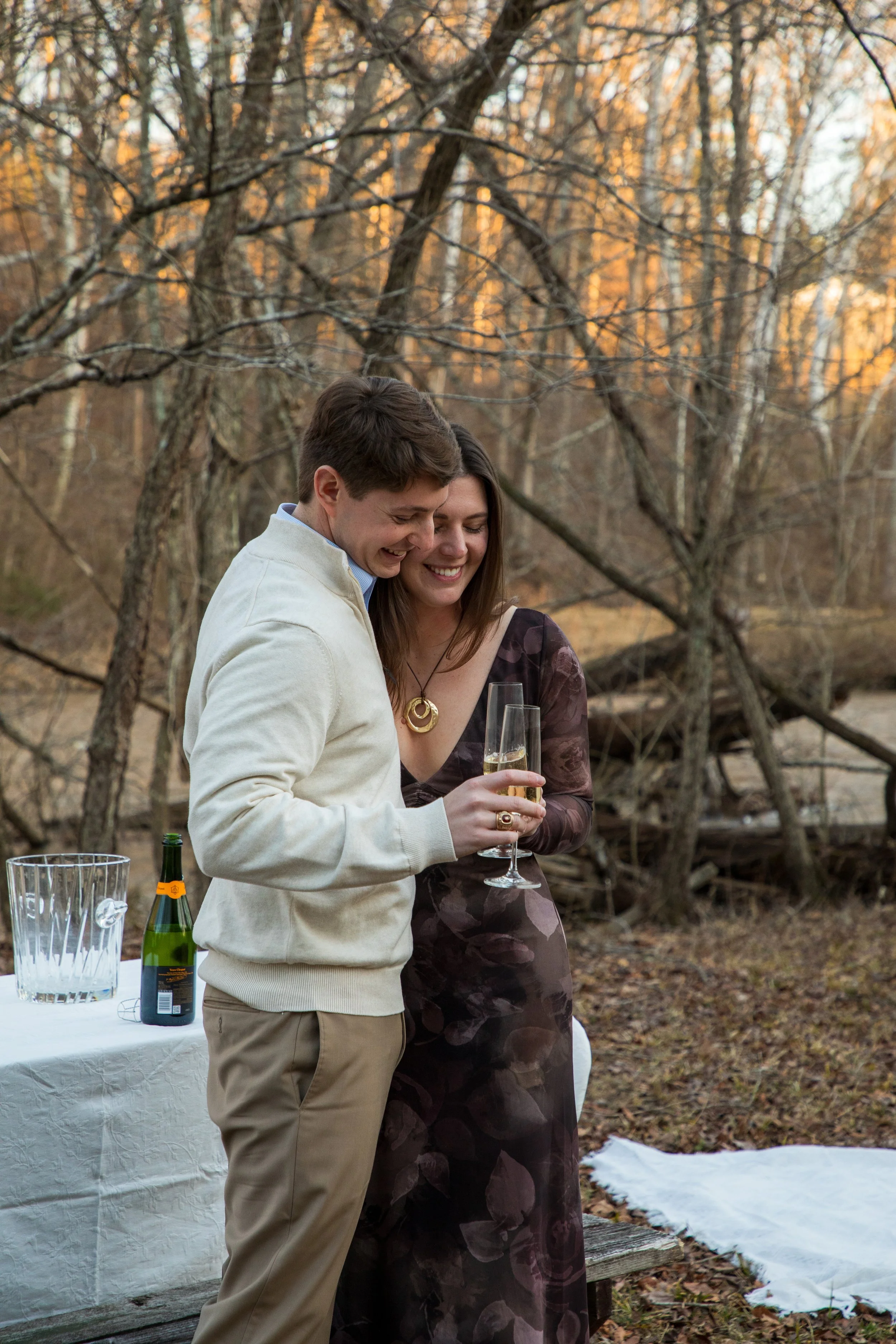 Engagement Photography by Will Locke near Richmond, VA in Montpelier. A man and woman smiling and hugging while holding champagne glasses outdoors during sunset, with a table of flowers nearby.