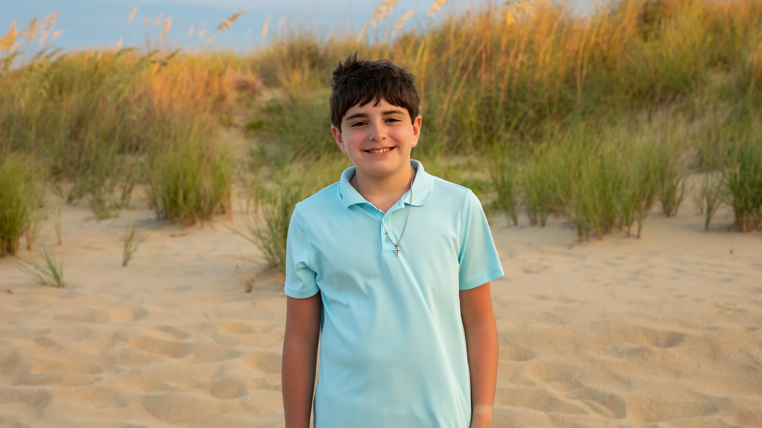 Photo & Video by Will Locke Family Photography in Virginia Beach, VA. A young boy with dark hair wearing a light blue polo shirt and a cross necklace, standing on a sandy beach with grass dunes and a blue sky in the background.