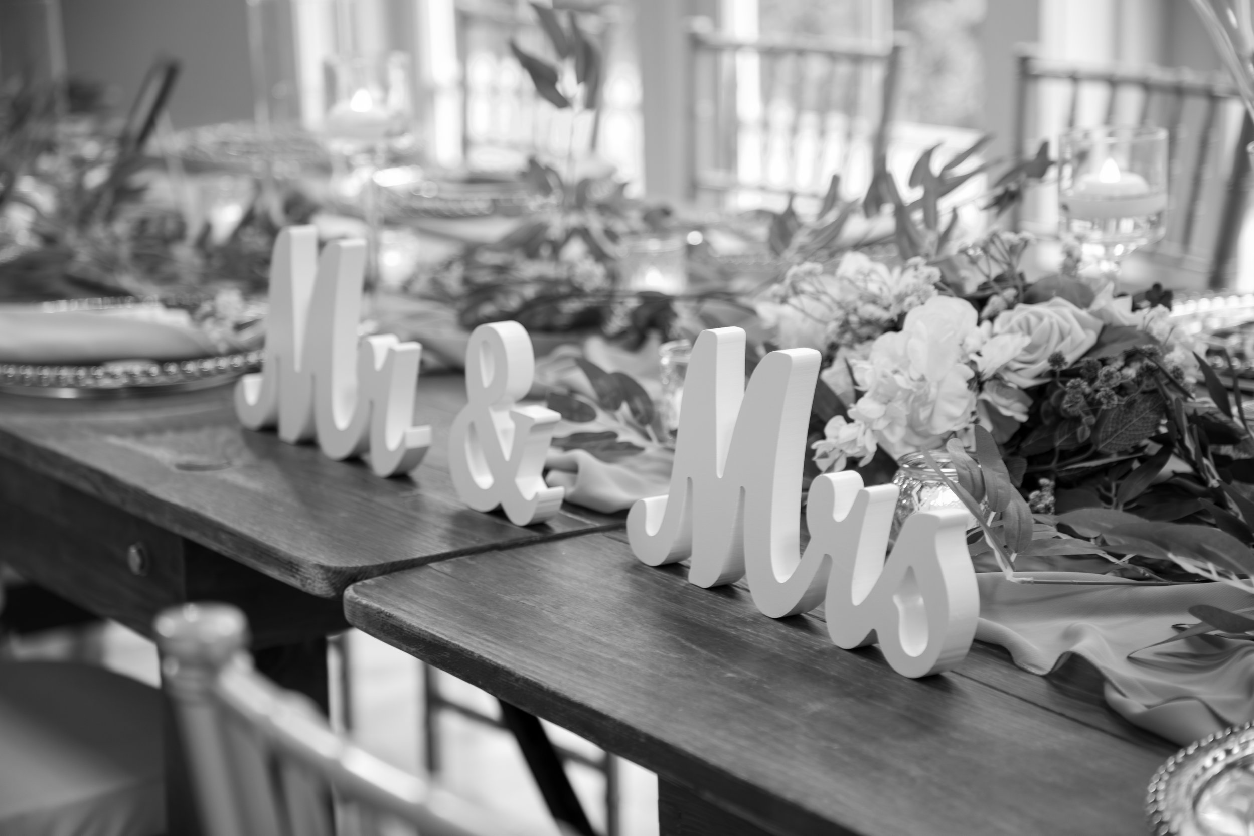 Wedding Photography by Will Locke. Decorative wedding table centerpiece with white floral arrangements and a sign that reads 'Mr & Mrs' on a wooden table in a well-lit room.