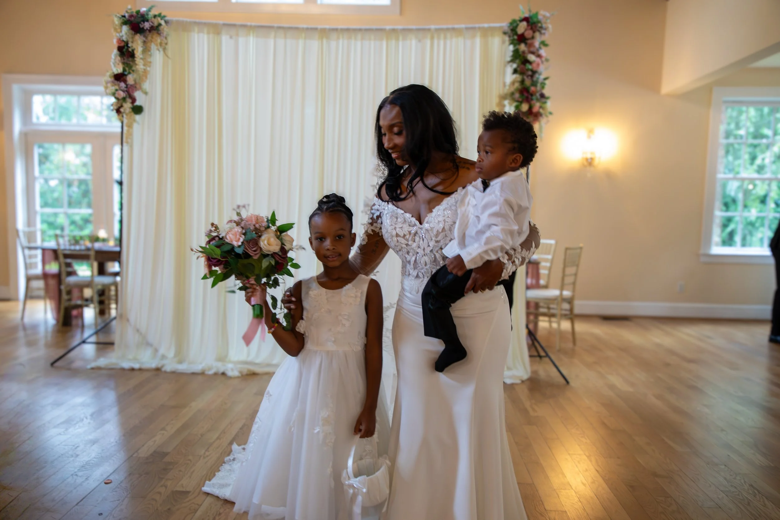Photo & Video by Will Locke Wedding Photography at the Woman's Club of Portsmouth in VA. A bride holding a young boy, a young girl holding a bouquet, all dressed in white, in a decorated indoor venue with floral arrangements and curtains.