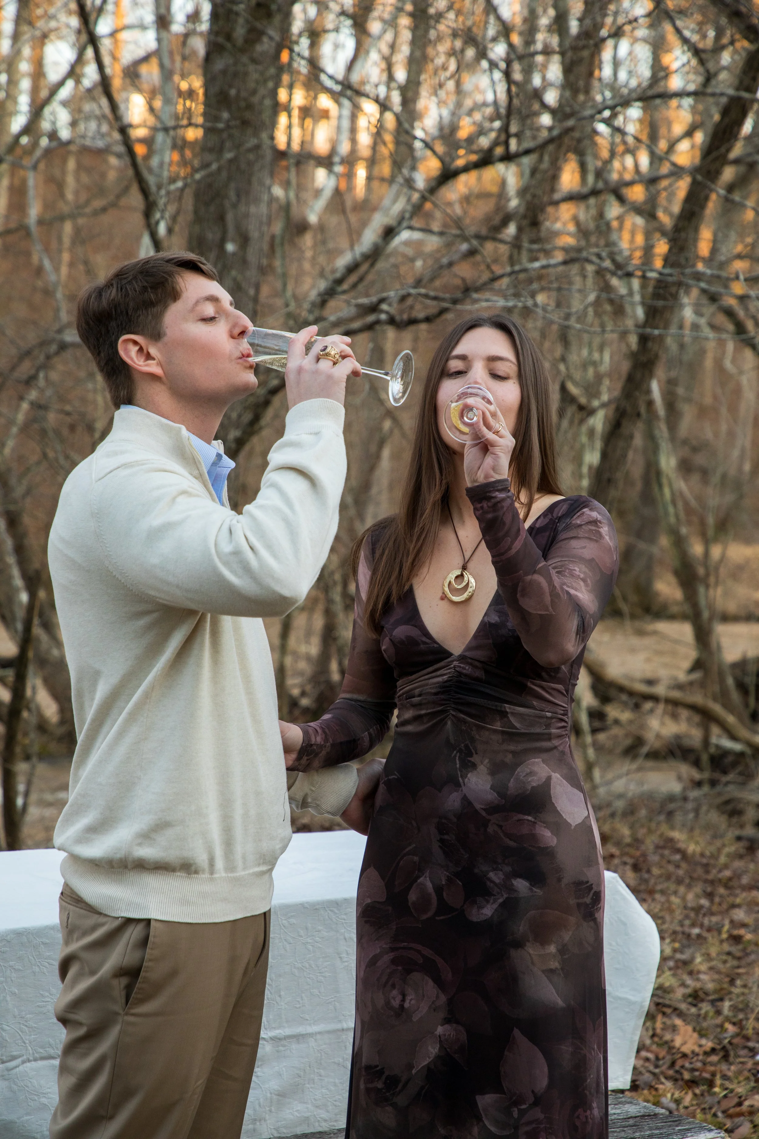 Engagement Photography by Will Locke near Richmond, VA in Montpelier. A couple sips champagne while celebrating their engagement with trees and a river in the background.