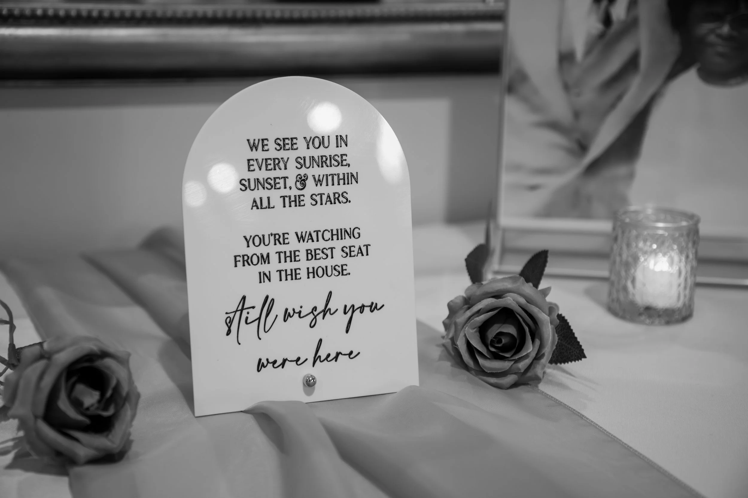 Wedding Photography by Will Locke. A white, arched table sign with a quote and rose decorations on a table, alongside a lit candle and a framed photo in the background.