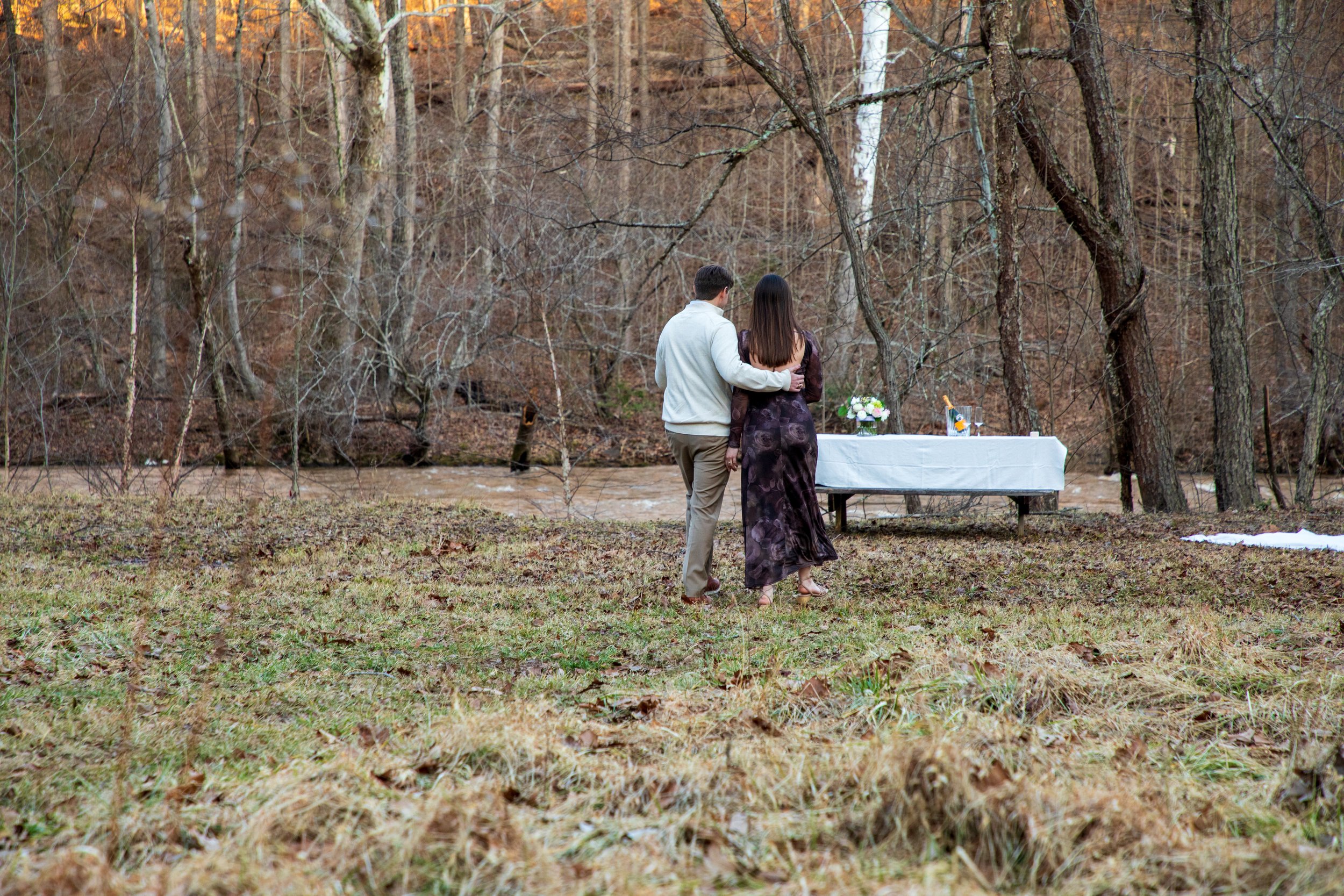 Engagement Photography by Will Locke near Richmond, VA in Montpelier. A couple walks to a white cloth on the ground near a wooded area with a stream on a fall day, with a table on the left side with white tablecloth.