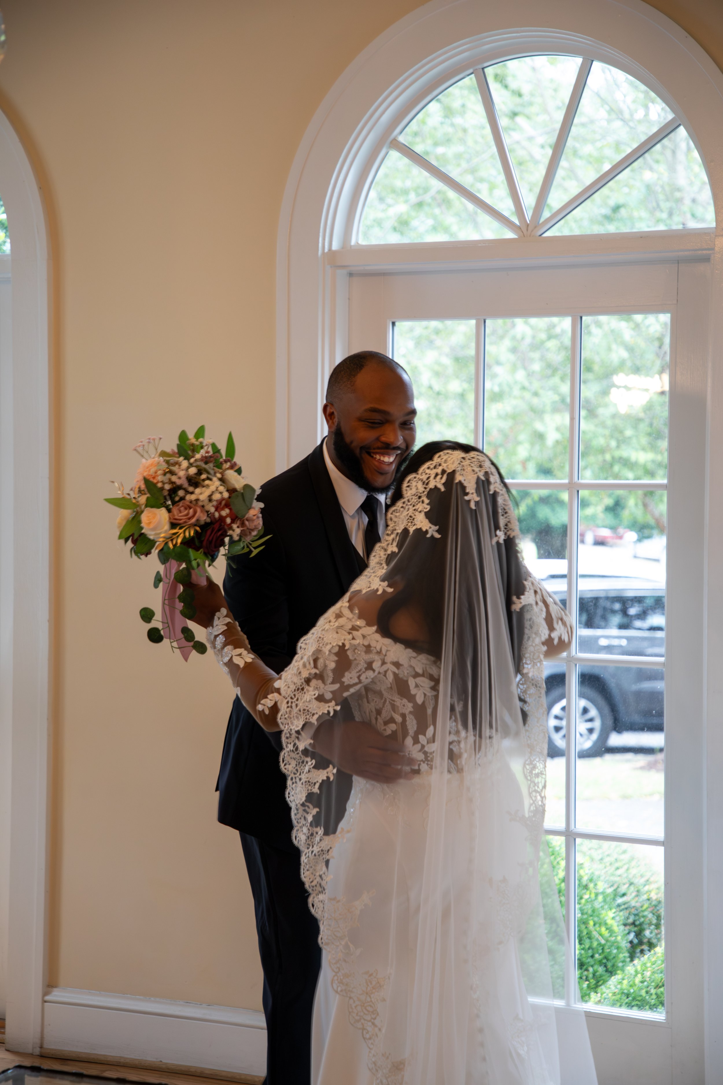 Wedding Photography by Will Locke. A couple in wedding attire sharing a joyful moment indoors near a window, with the bride holding a bouquet of flowers.