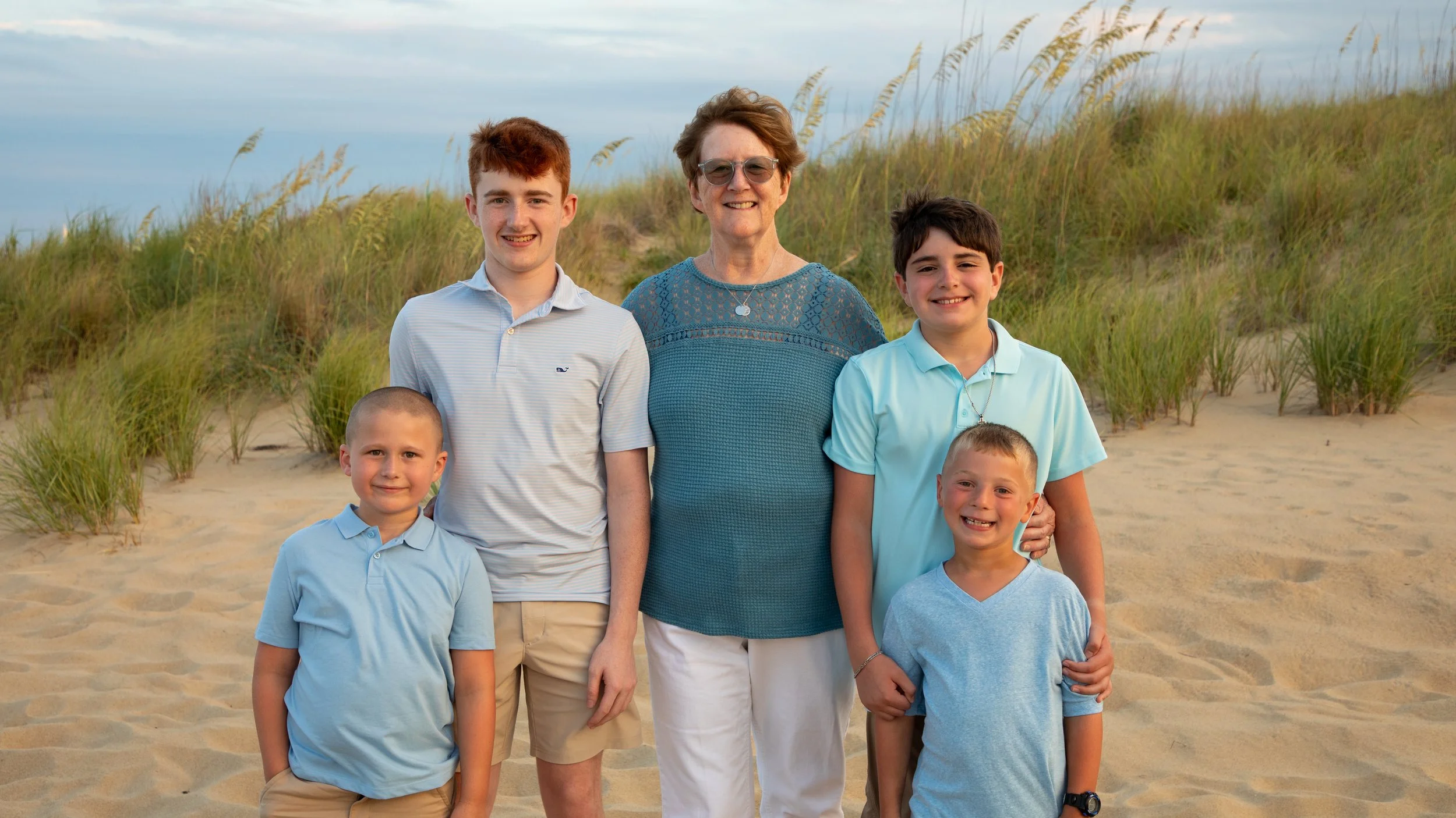 Photo & Video by Will Locke Family Photography in Virginia Beach, VA. Family of six people standing together on a sandy beach with grassy dunes in the background, smiling at the camera.