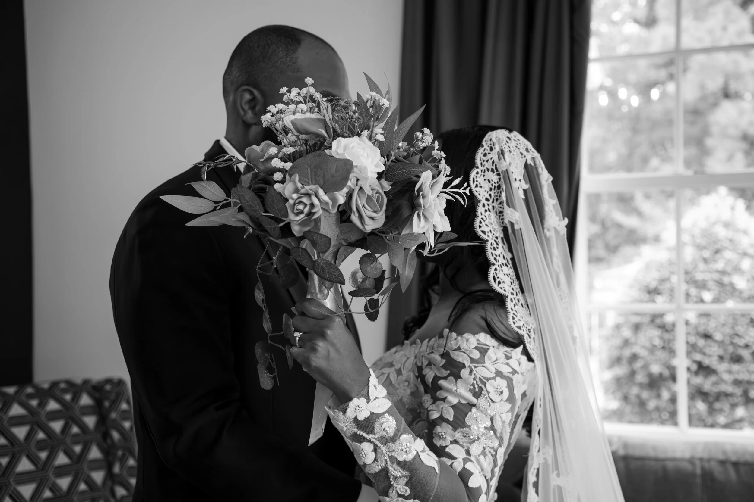 Wedding Photography by Will Locke. A black and white photo of a bride and groom with the bride holding a large bouquet covering their faces, in front of a window with curtains.