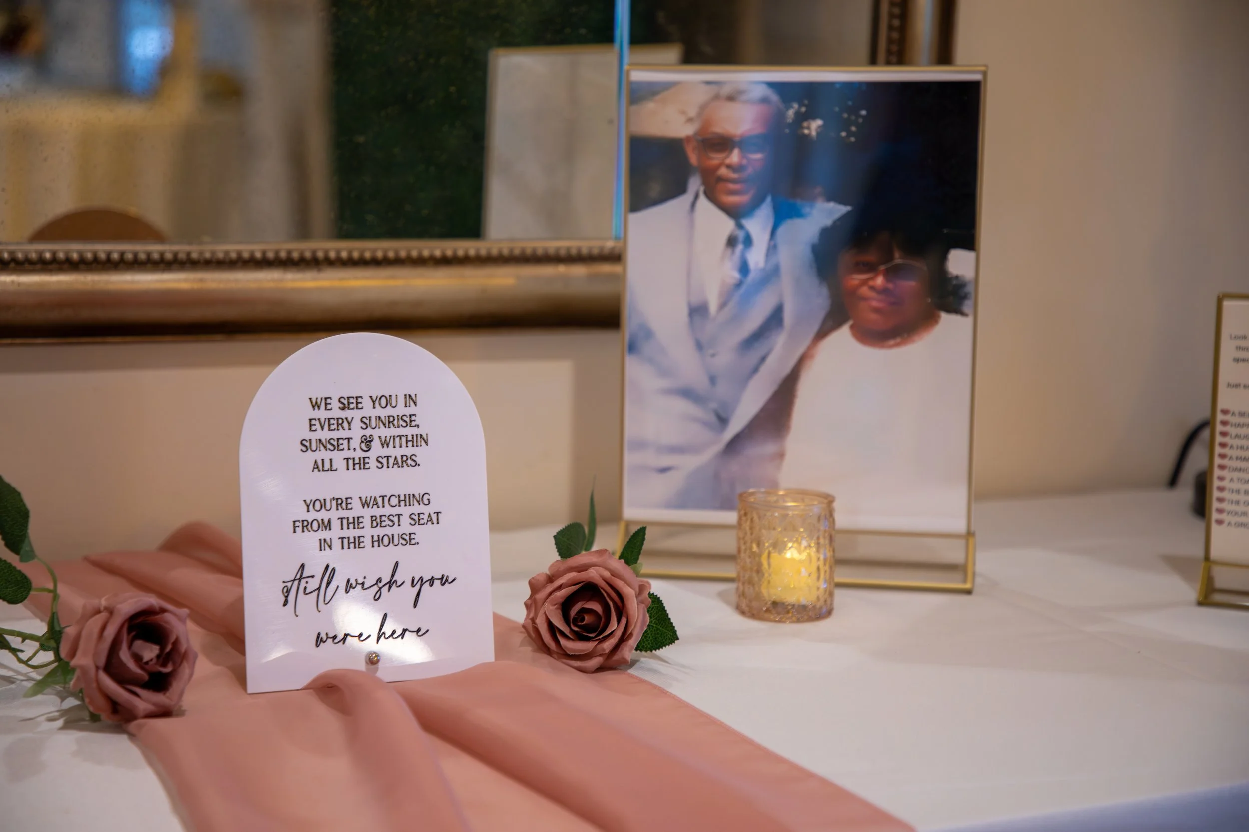 Wedding Photography by Will Locke. A memorial display with a framed photograph of an elderly couple, a candle in a glass holder, pink roses, a pink cloth, and a heartfelt message on a white paper