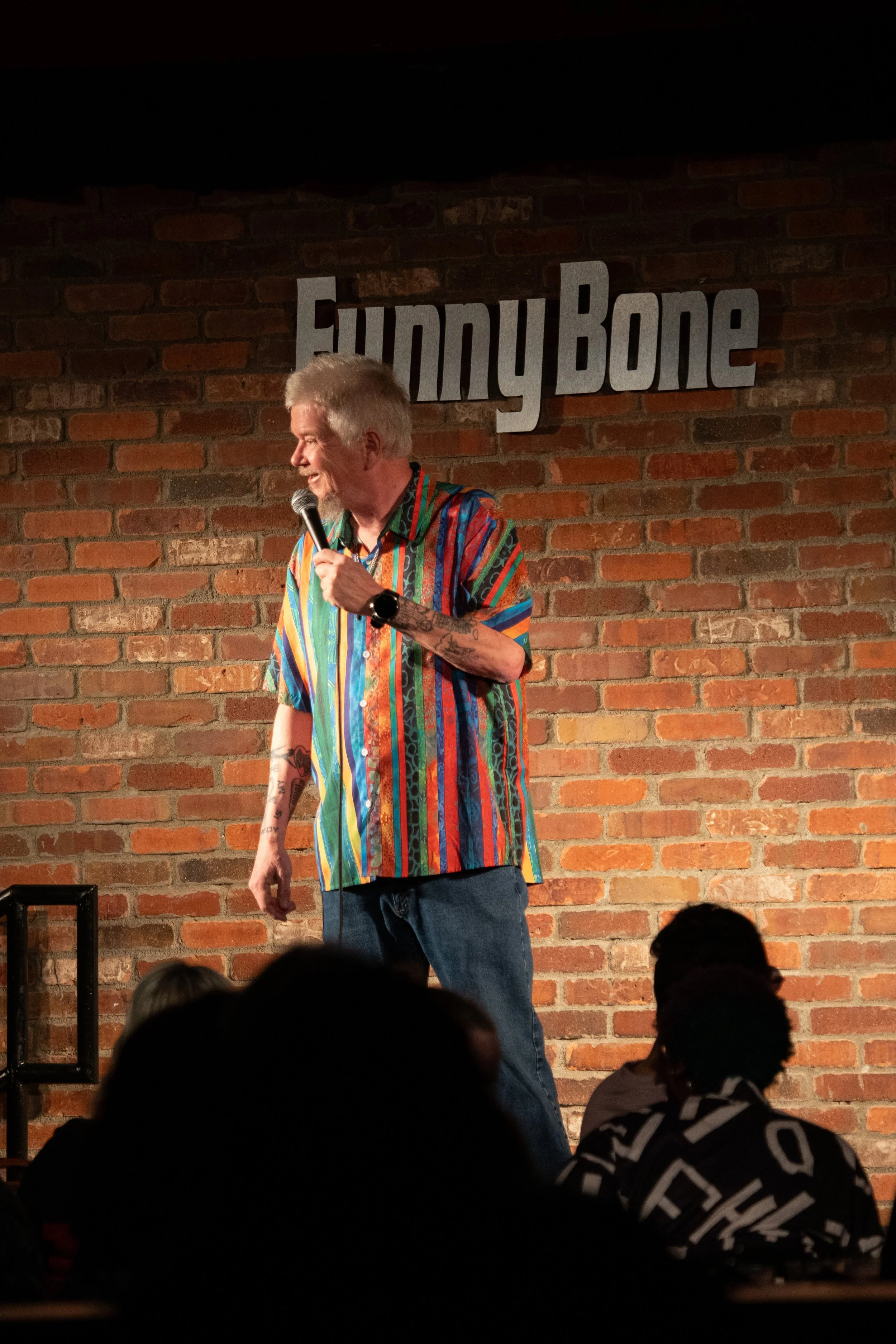 Comedy Photography by Will Locke. A comedian performs on stage at a comedy club called Funny Bone, with a brick wall background and audience members in front.