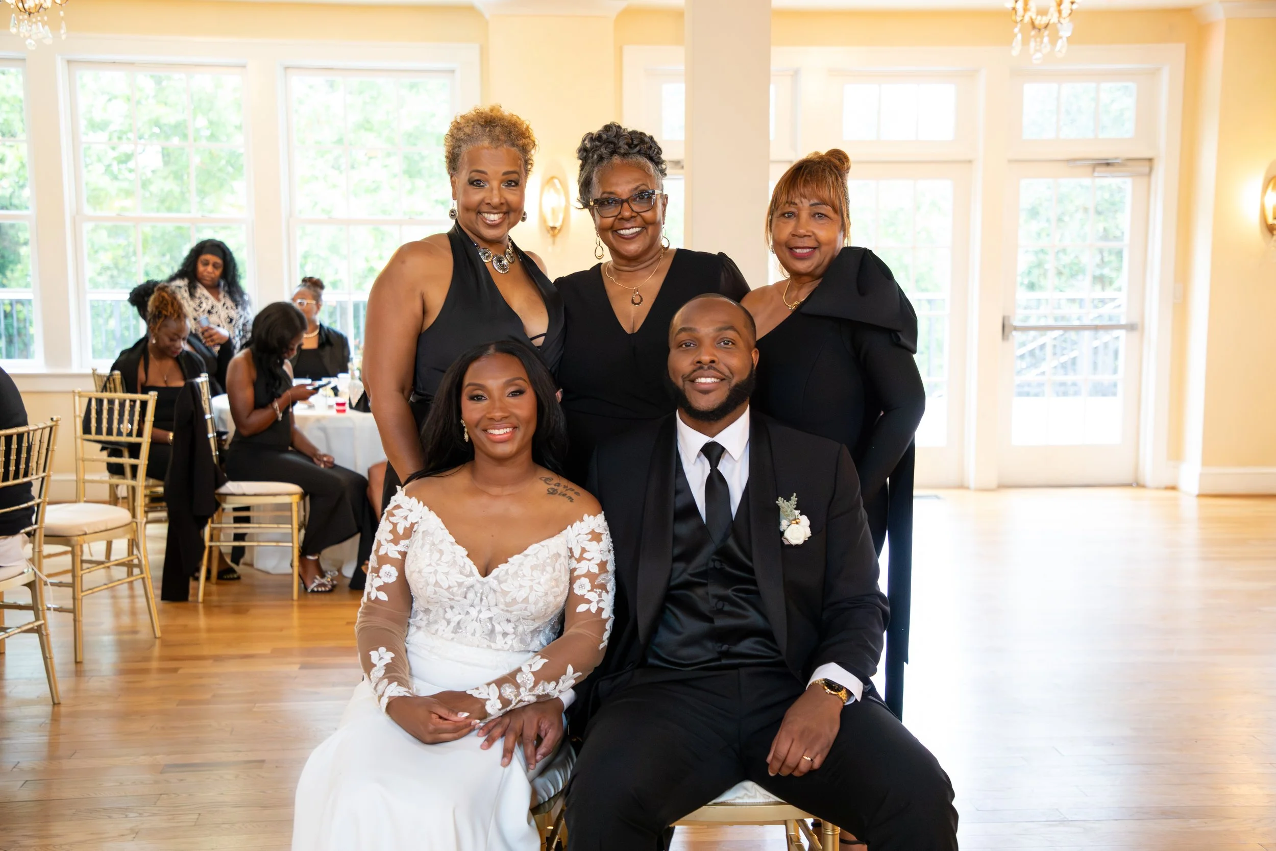 Photo & Video by Will Locke Wedding Photography at the Woman's Club of Portsmouth. Group of five people, including a bride in a white wedding dress and groom in a black suit, posing for a photo at a wedding reception with guests in the background.