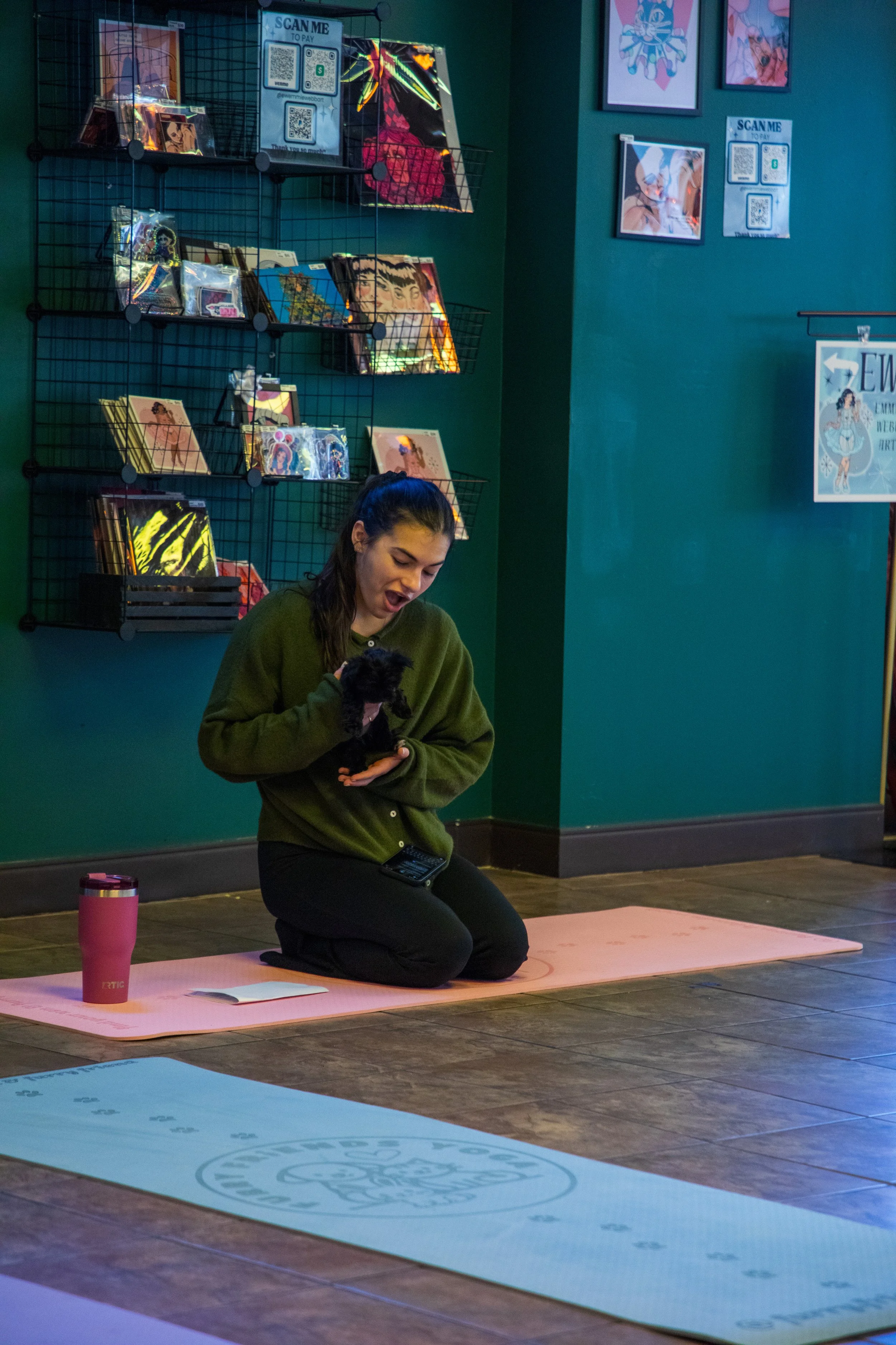 A woman kneeling on a pink yoga mat, holding a small black puppy, with a pink travel mug, notebook, and remote control nearby. Marketing and Event Photography by Will Locke.