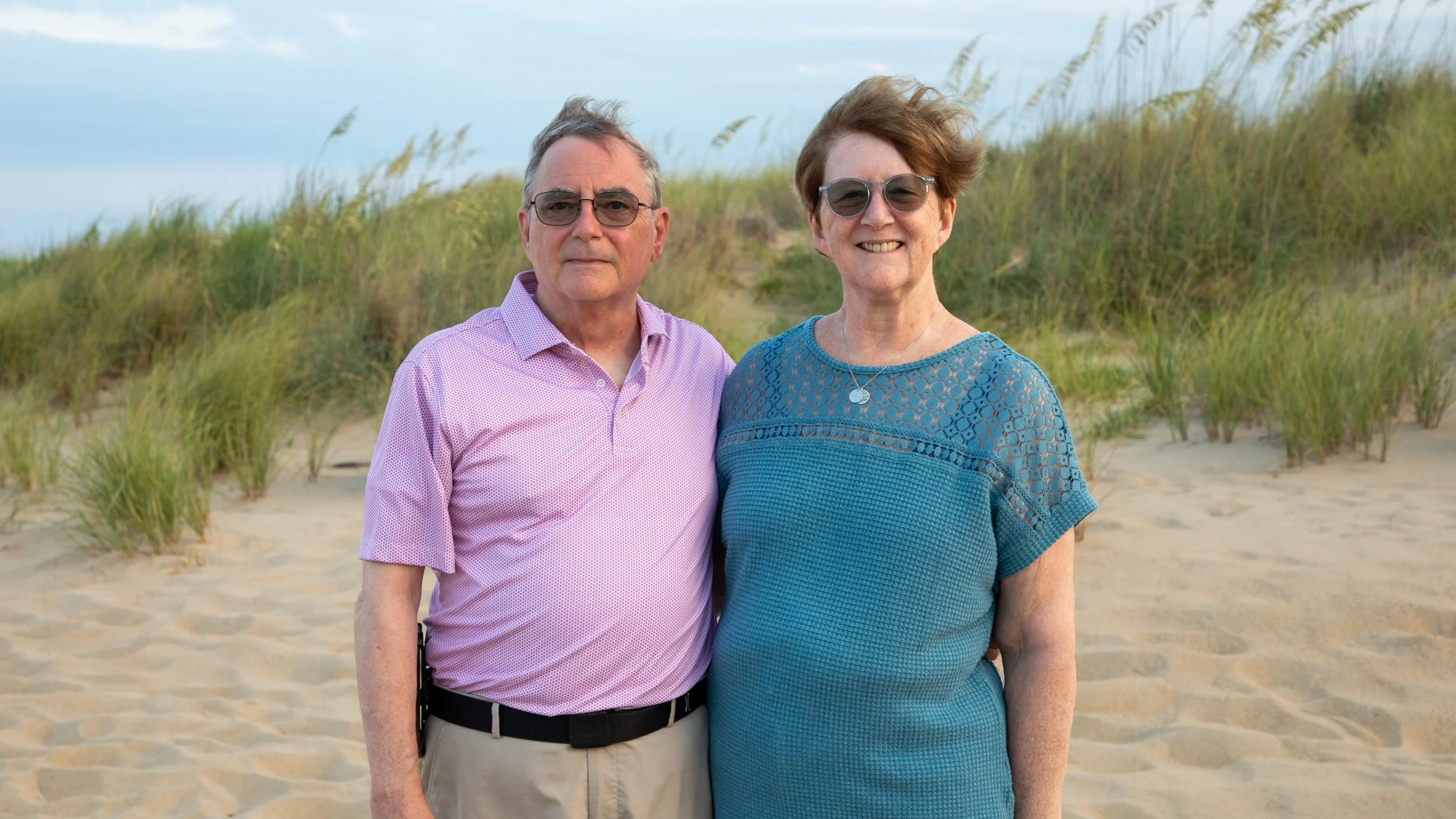 Photo & Video by Will Locke Family Photography in Virginia Beach, VA. An elderly man and woman standing on a sandy beach with green sand dunes and grass in the background, smiling and wearing sunglasses.