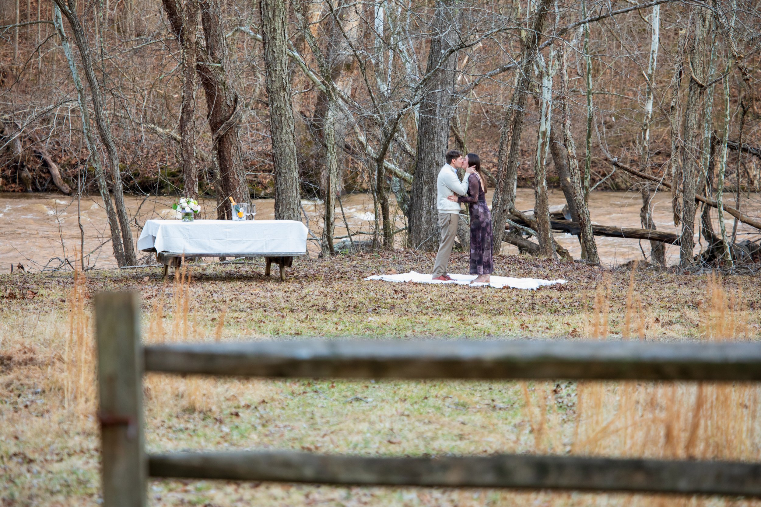 Engagement Photography by Will Locke near Richmond, VA in Montpelier. A couple kisses each other outdoors near a river, standing on a white blanket on the ground, surrounded by leafless trees in a forested area.