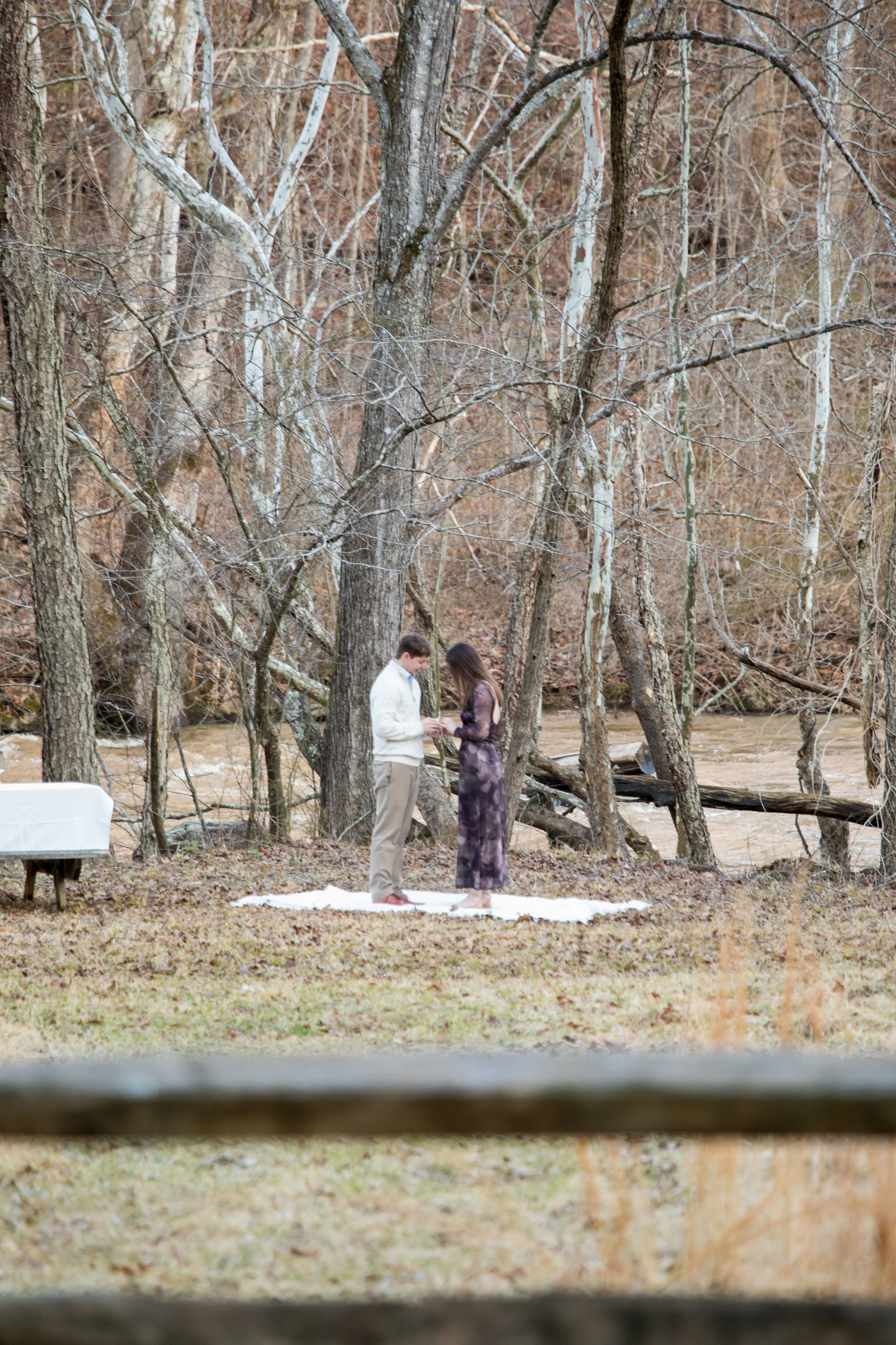 Engagement Photography by Will Locke near Richmond, VA in Montpelier. A couple standing on a white cloth outdoors by a wooded area with leafless trees and a river in the background, participating in an outdoor ceremony.