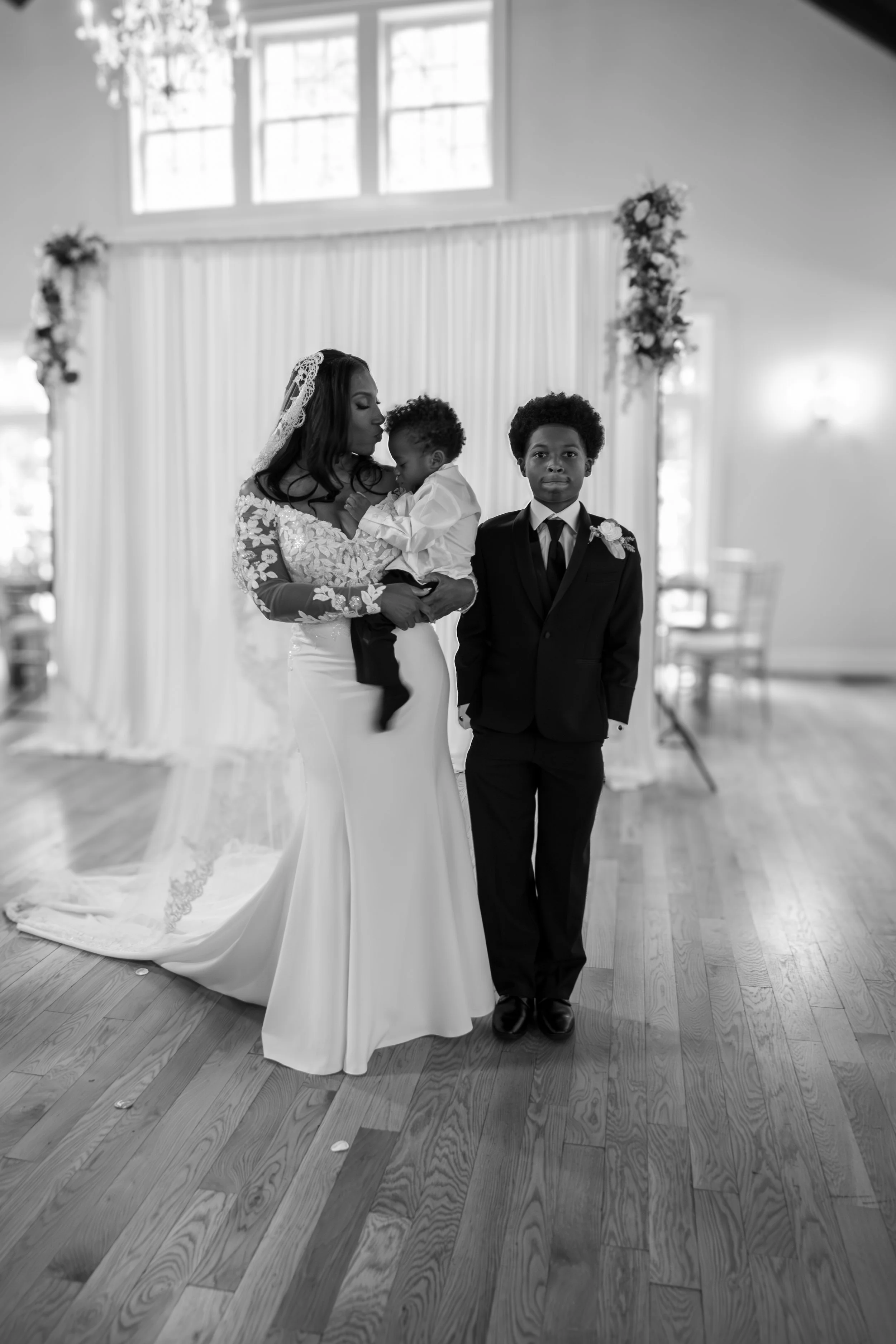 Photo & Video by Will Locke Wedding Photography at the Woman's Club of Portsmouth in VA. Woman in a wedding dress holds a small child, while a young boy in a suit stands beside her in a decorated indoor setting with wooden flooring and large windows.