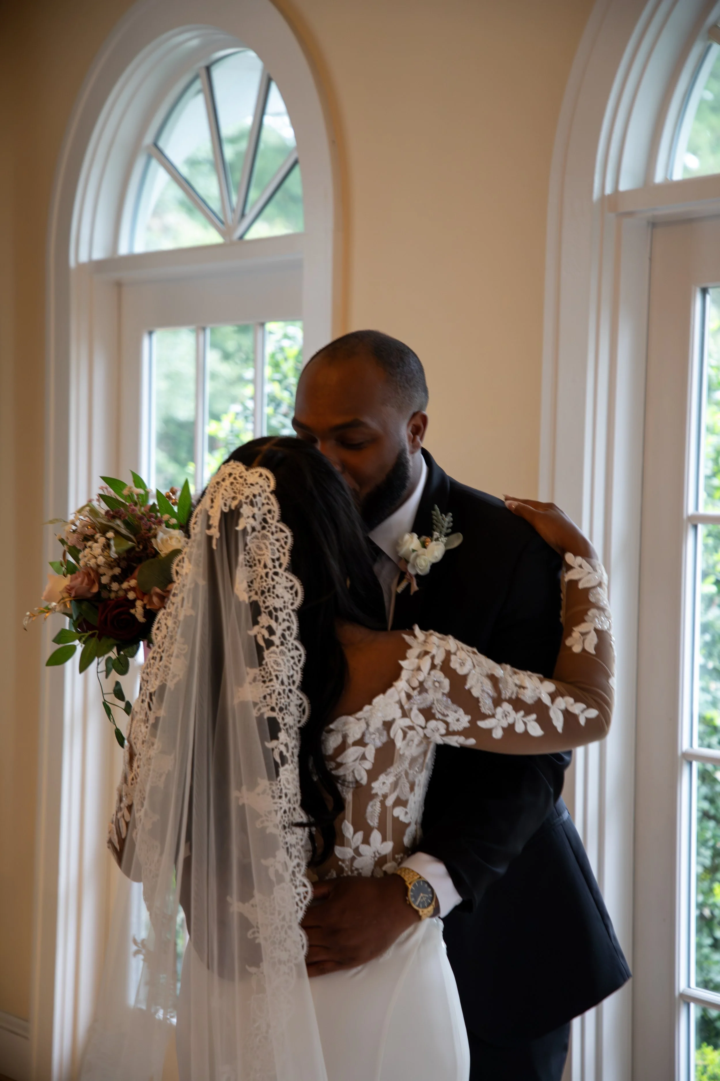 Photo & Video by Will Locke Wedding Photography at the Woman's Club of Portsmouth in VA. A bride and groom share a kiss indoors near large windows, with the bride wearing a lace wedding dress and veil holding a bouquet, and the groom in a dark suit.