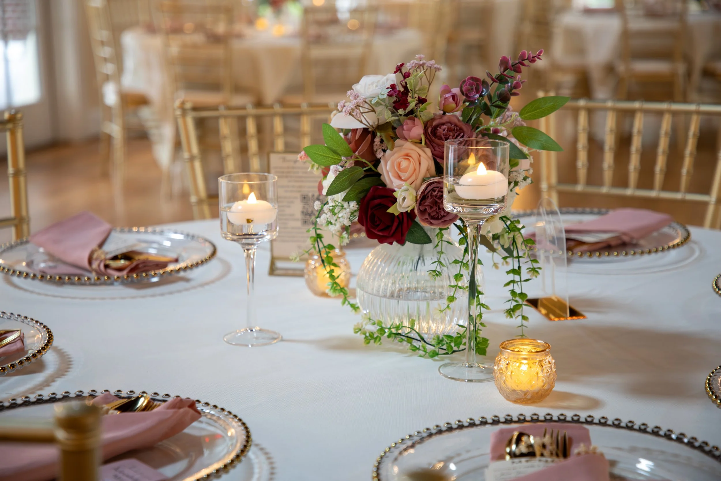 Wedding Photography by Will Locke. Elegant wedding reception table with a floral centerpiece, candles, and place settings with pink napkins on gold-trimmed plates.