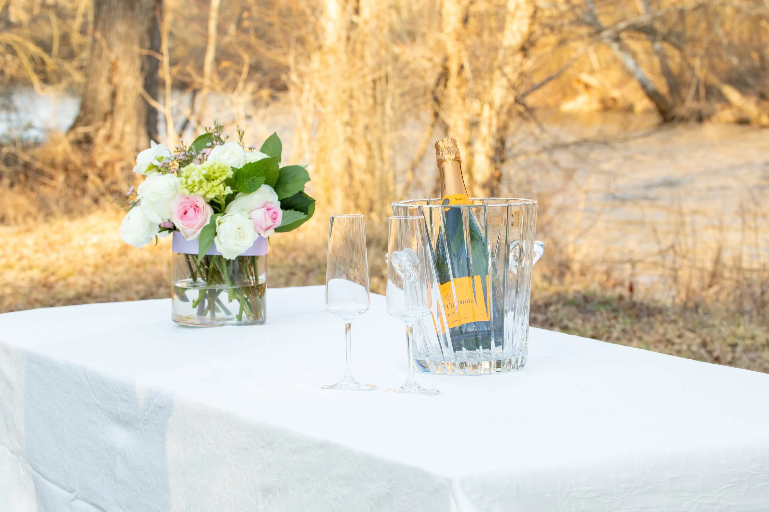Engagement Photography by Will Locke near Richmond, VA in Montpelier. A bottle of Veuve Clicquot champagne in an ice bucket with two empty champagne flutes on a white table outdoors, with a flower arrangement. 