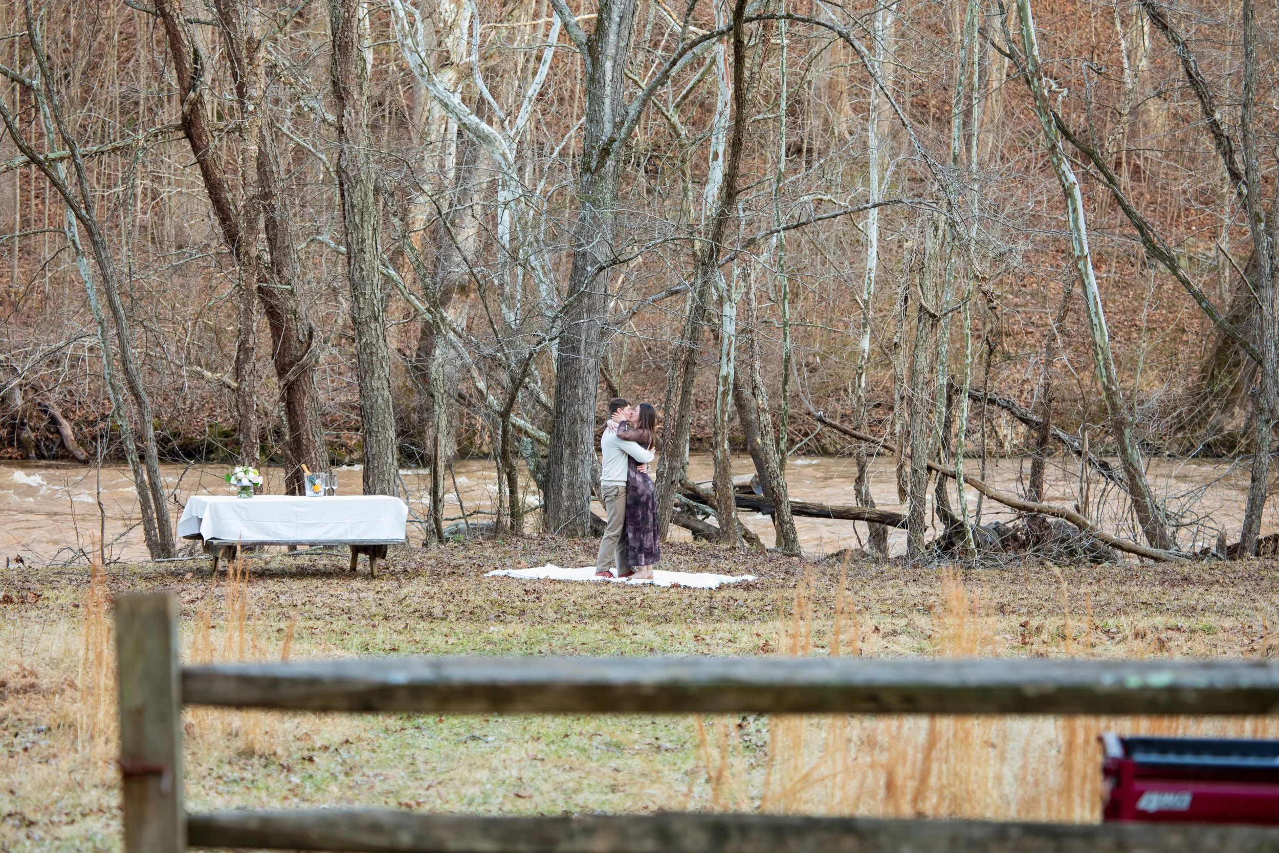 Engagement Photography by Will Locke near Richmond, VA in Montpelier. A couple stands hugging and kissing in a wooded outdoor setting near a river, with a white table with flowers and a bottle in the background.