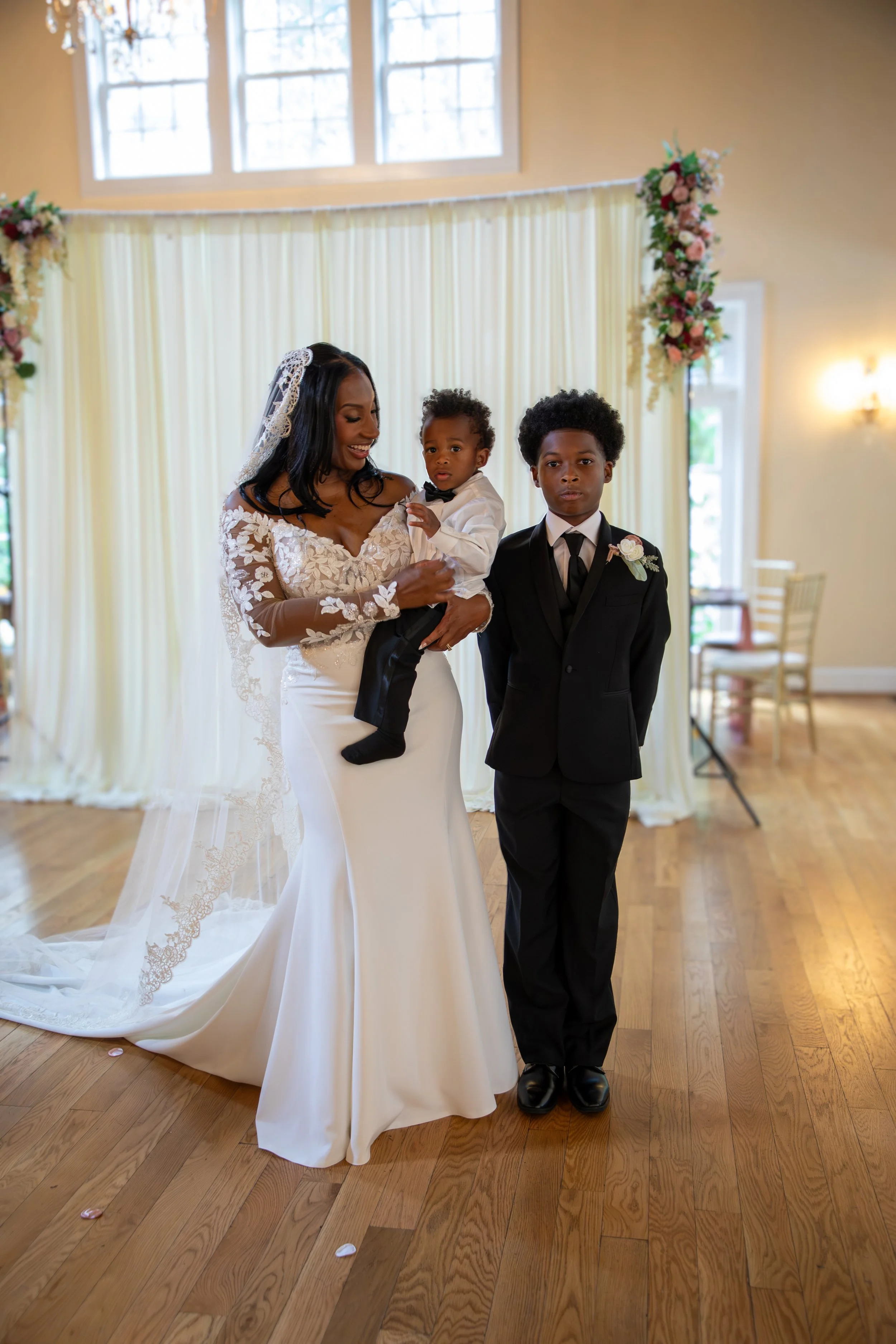 Photo & Video by Will Locke Wedding Photography at the Woman's Club of Portsmouth. A bride in a white wedding dress holding a young boy in a tuxedo, standing next to a young man in a black tuxedo at a wedding ceremony indoors with floral decorations.