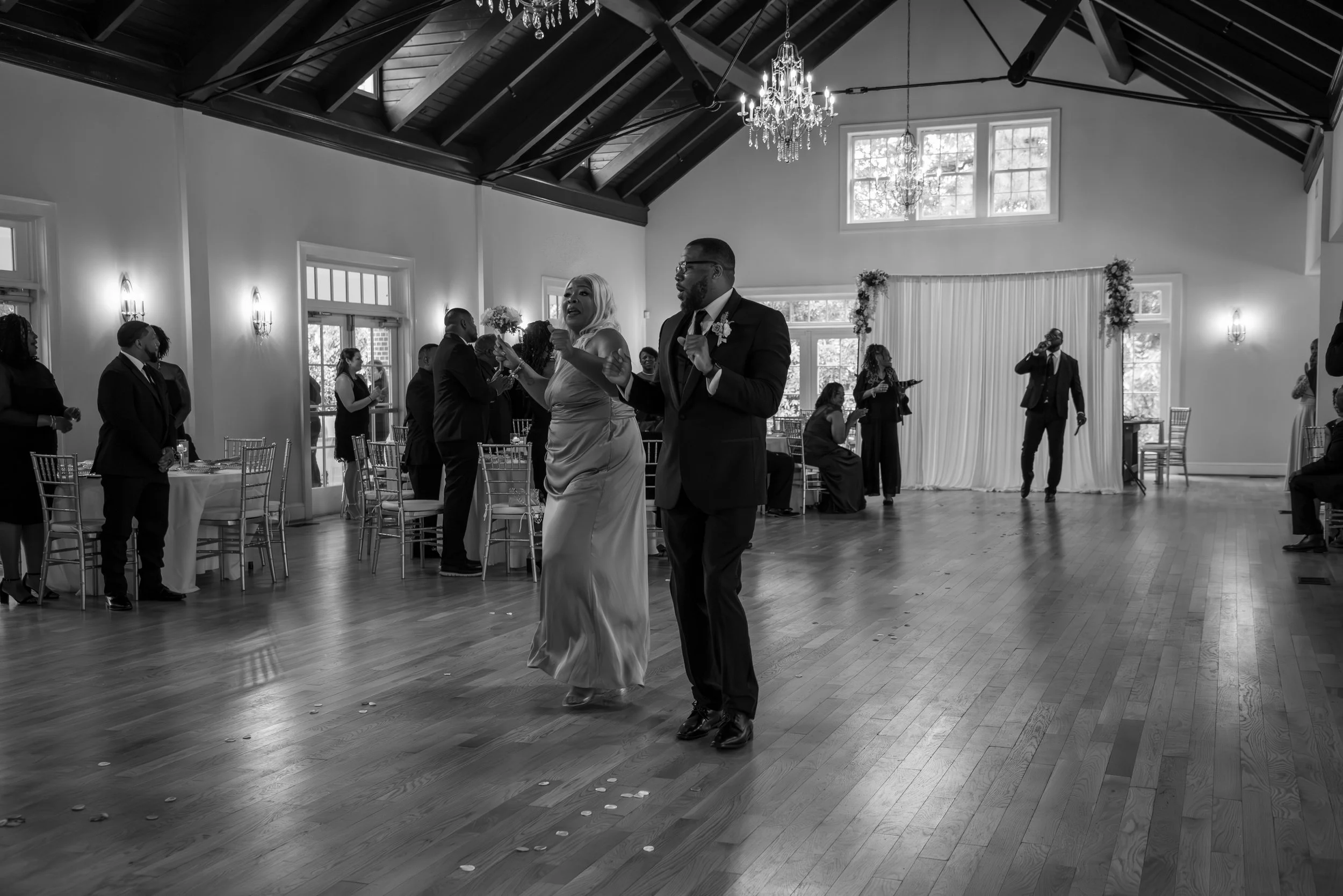 Wedding Photography by Will Locke. A couple dancing at a wedding reception in a spacious hall with high vaulted ceilings, chandeliers, and large windows, surrounded by guests watching.