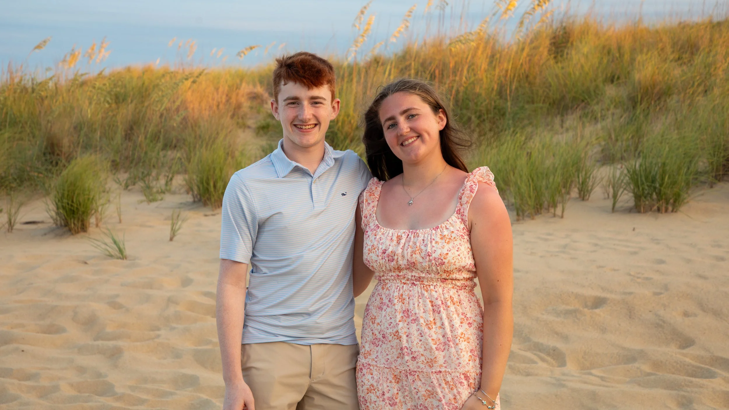 Photo & Video by Will Locke Family Photography in Sandbridge, Virginia Beach, VA at Little Island Park. A young man and woman standing on a sandy beach with grass-covered dunes in the background, smiling at the camera during sunset.