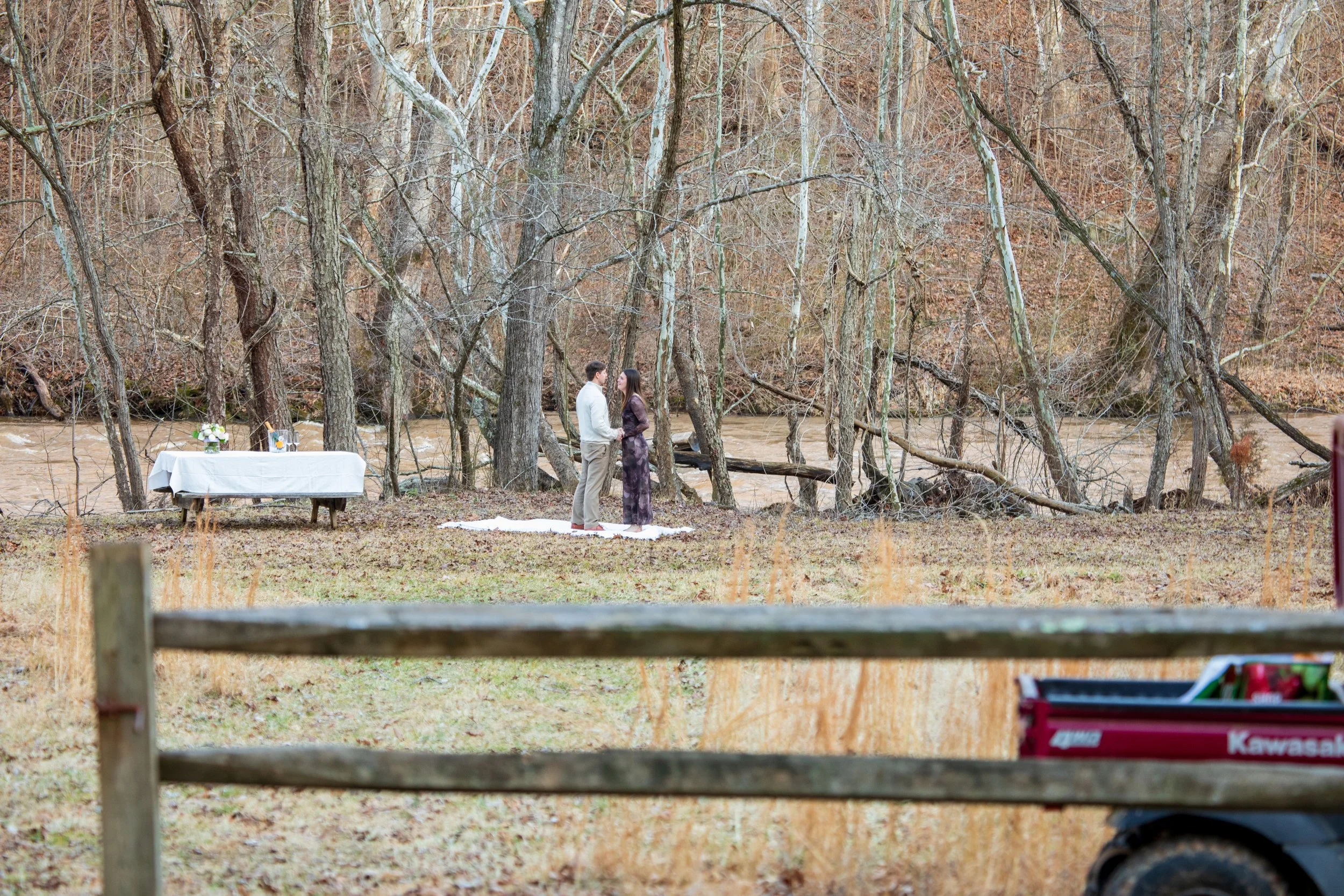 Engagement Photography by Will Locke near Richmond, VA in Montpelier. A couple walks on a white cloth on the ground near a table with tablecloth in a wooded area with a stream on a fall day, with a Kawasaki utility vehicle.