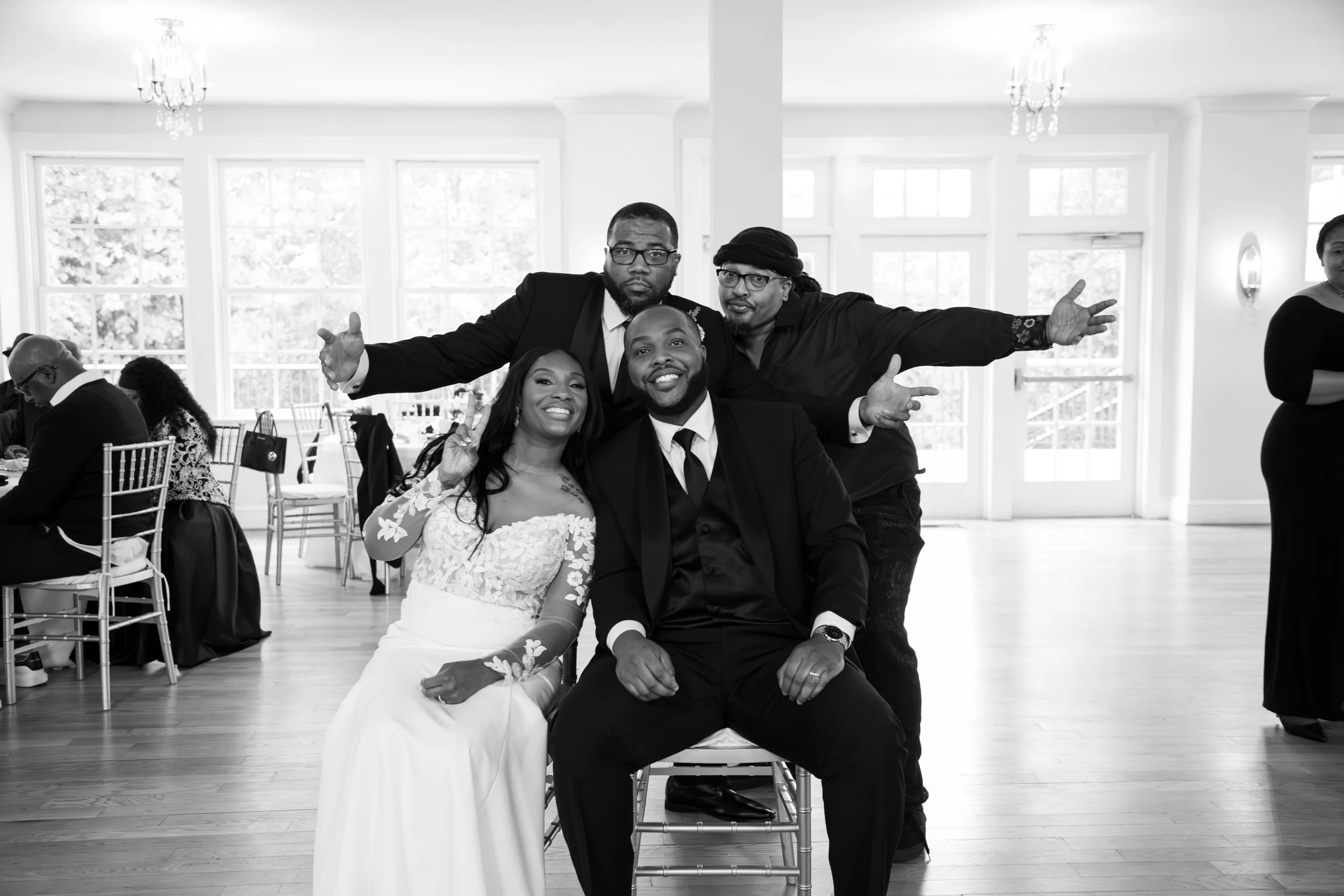 Photo & Video by Will Locke Wedding Photography at the Woman's Club of Portsmouth in Virginia. A group of four people at a celebratory event, with the woman in a white lace dress sitting and the man in a suit sitting on a chair.