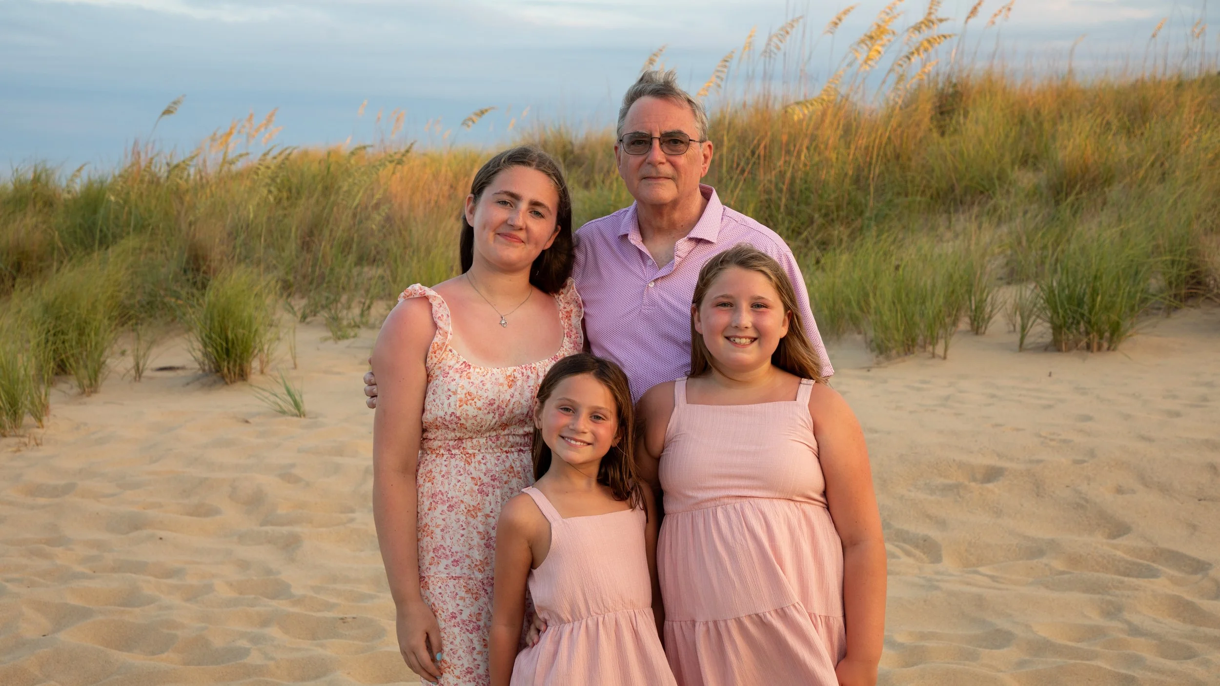 A family of five people standing on a sandy beach with tall grass and blue sky in the background. Photo & Video by Will Locke Family Photography in Virginia Beach, VA. 