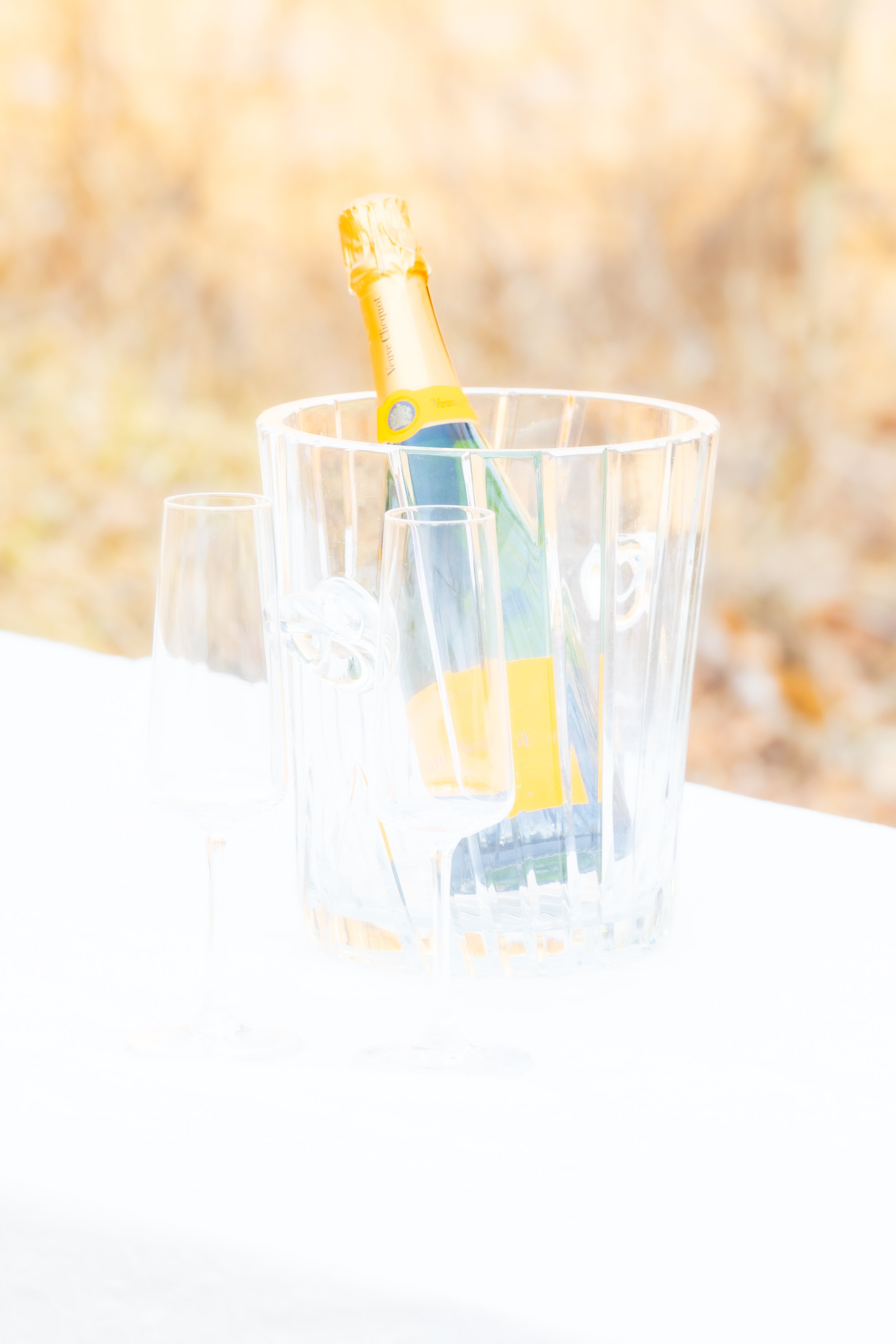 Engagement Photography by Will Locke near Richmond, VA in Montpelier. A bottle of Veuve Clicquot champagne in an ice bucket with two empty champagne flutes on a white table outdoors, with a flower arrangement. 