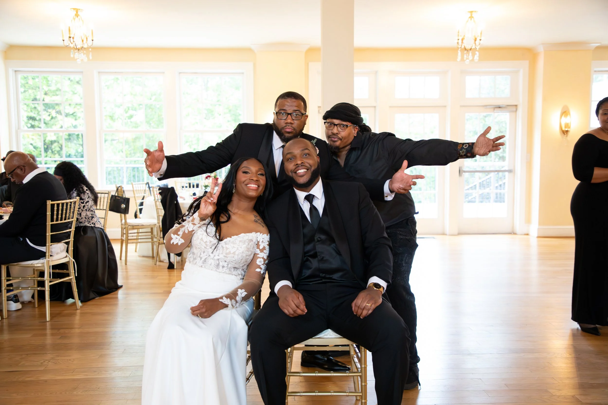 Photo & Video by Will Locke Wedding Photography at the Woman's Club of Portsmouth in Virginia. Group of four people, two men and two women, posing and smiling at a wedding reception indoor venue with large windows and chandeliers.