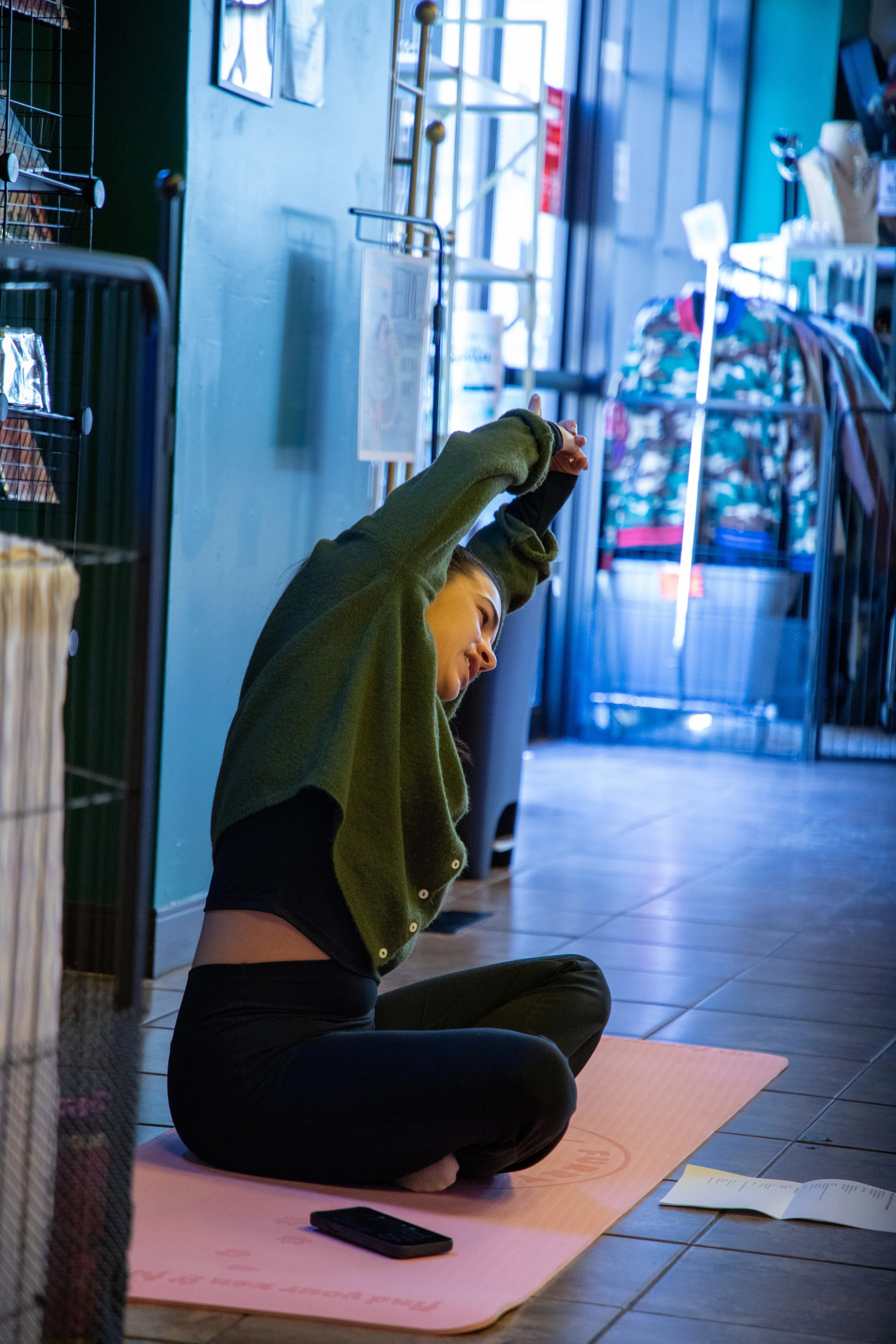 Marketing and Event Photography by Will Locke. A woman sitting on a pink yoga mat in a store, stretching with her arms up, wearing black leggings and a loose green cardigan, with a cellphone and papers nearby.