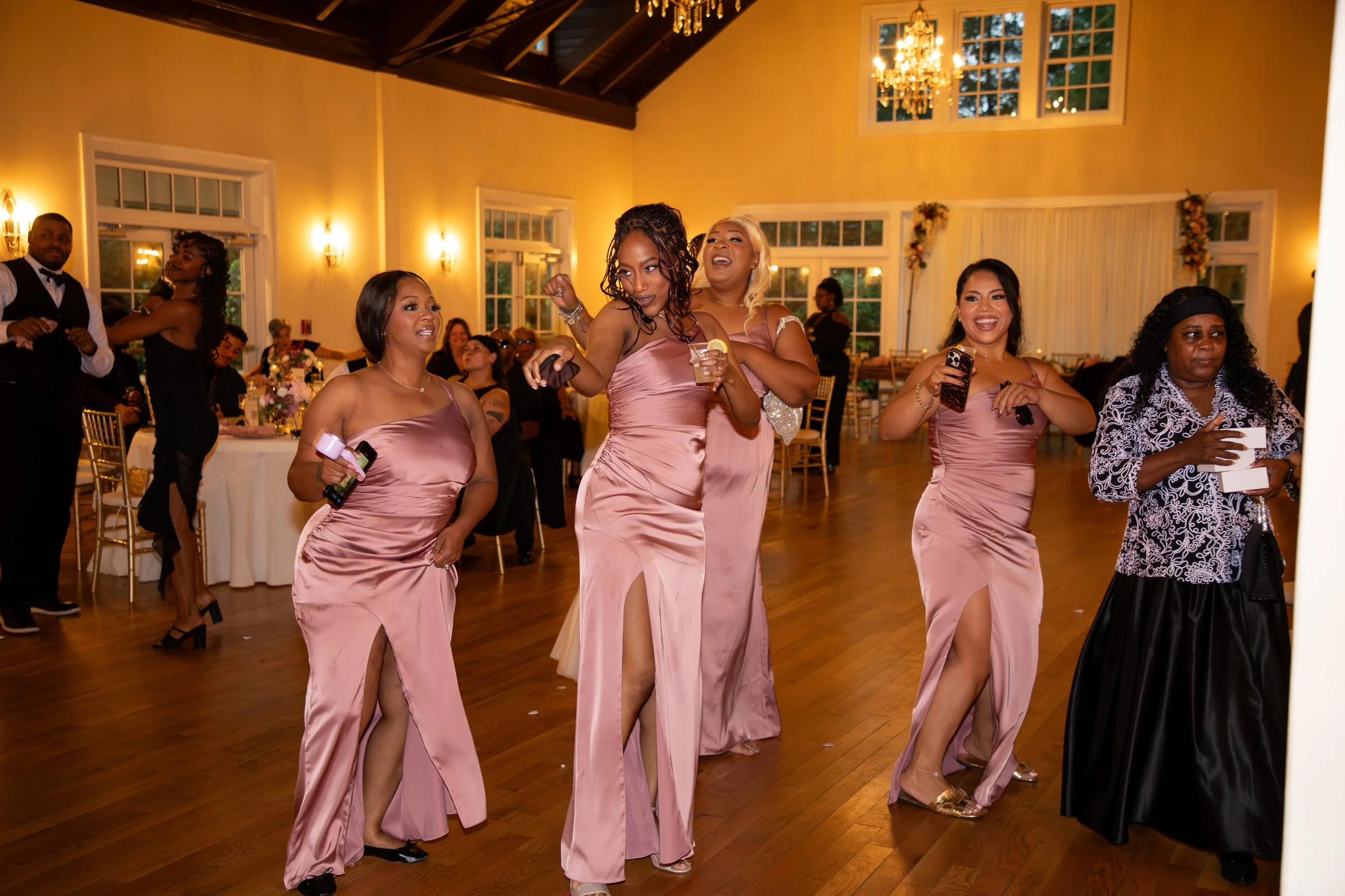 Wedding Photography by Will Locke. Group of women in pink satin dresses dancing and enjoying themselves at a wedding reception in a decorated hall with chandeliers and large windows.