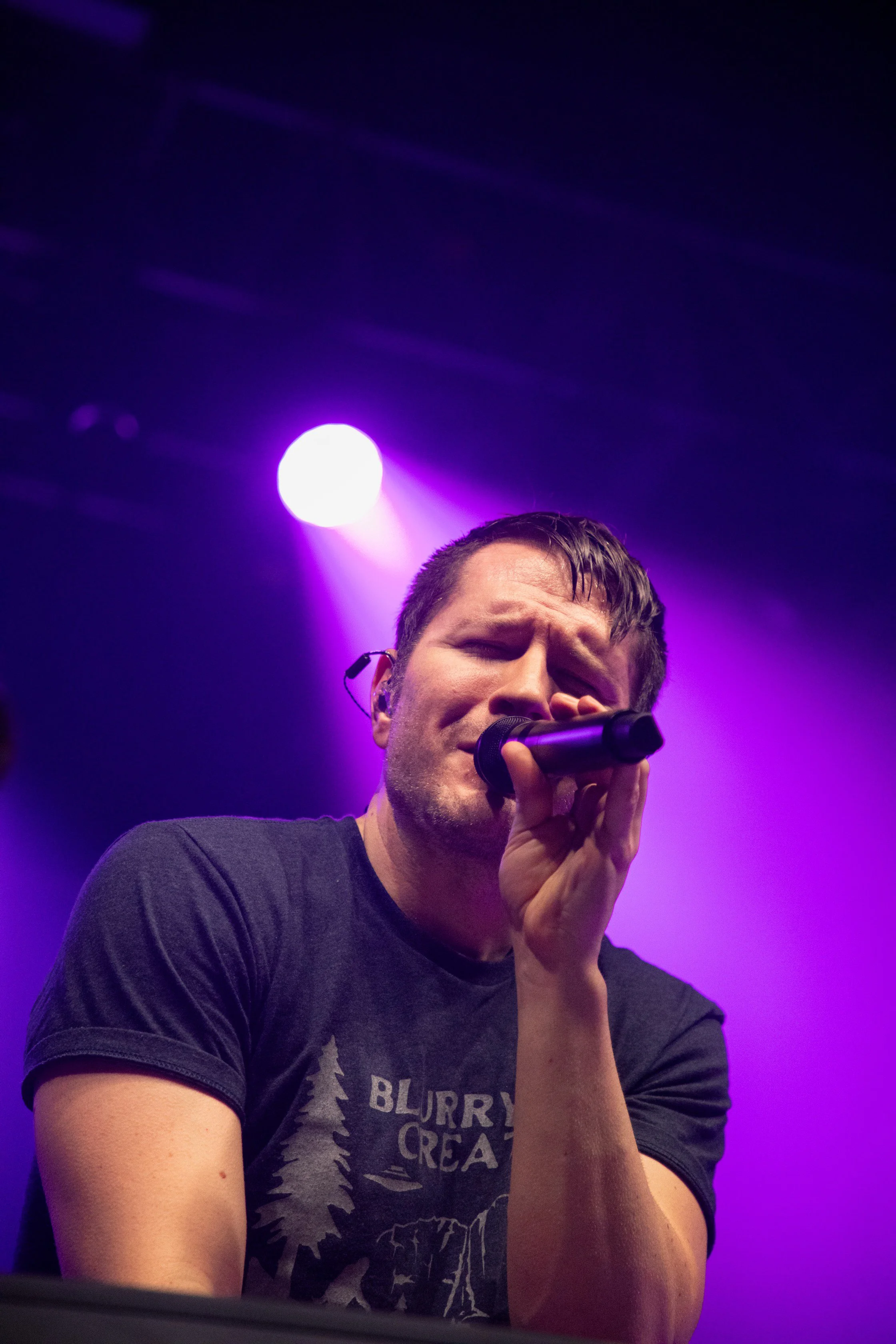 Concert Photography by Will Locke. A man singing into a microphone on stage with purple lighting in the background.